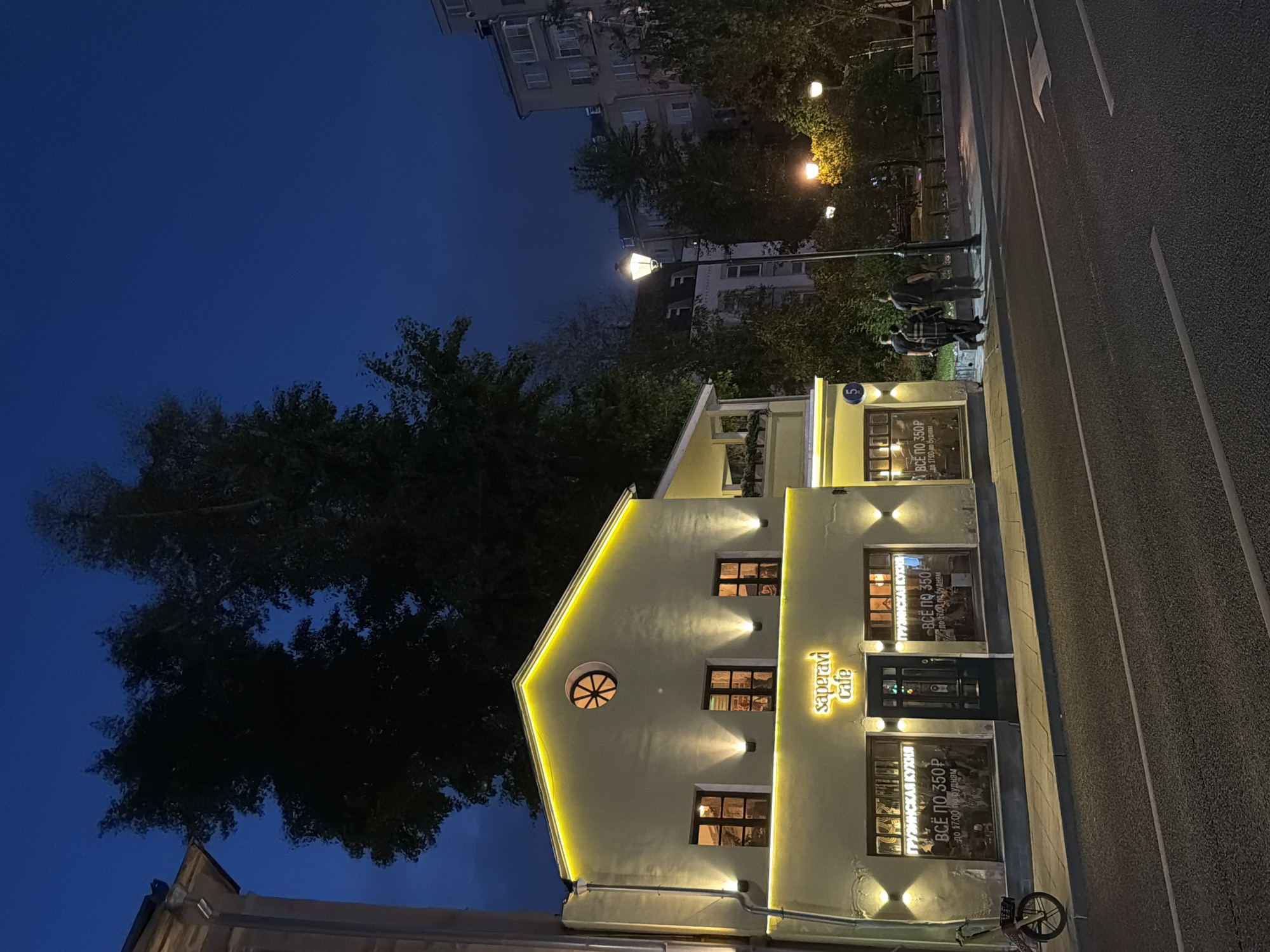 The image depicts a street scene at dusk or night in Moscow, Russia. In the foreground, a paved road with white markings runs diagonally from right to left. To the left of the road, a sidewalk is visible, bordering a light yellow building. This building has a pitched roof illuminated by yellow lighting along its edge and spotlights on its facade. It features several windows with dark frames, some of which are lit from within. A sign above the entrance reads "Saperavi cafe" in yellow lettering. Some windows display signs with Russian text, including "ГРУЗИНСКАЯ КУХНЯ" (Georgian Cuisine) and "ВСЁ ПО 350Р" (Everything for 350 rubles). A bicycle with a basket is parked on the sidewalk to the far left.
Further down the sidewalk, three individuals are walking away from the camera. Two are men dressed in dark clothing, and one appears to be wearing a checkered shirt. A streetlamp with a single light source stands near them. Beyond the cafe and the streetlamp, trees and other buildings are visible, including a taller apartment building in the background. The sky is a deep, dark blue, suggesting late evening. The overall impression is a quiet street in an urban environment.