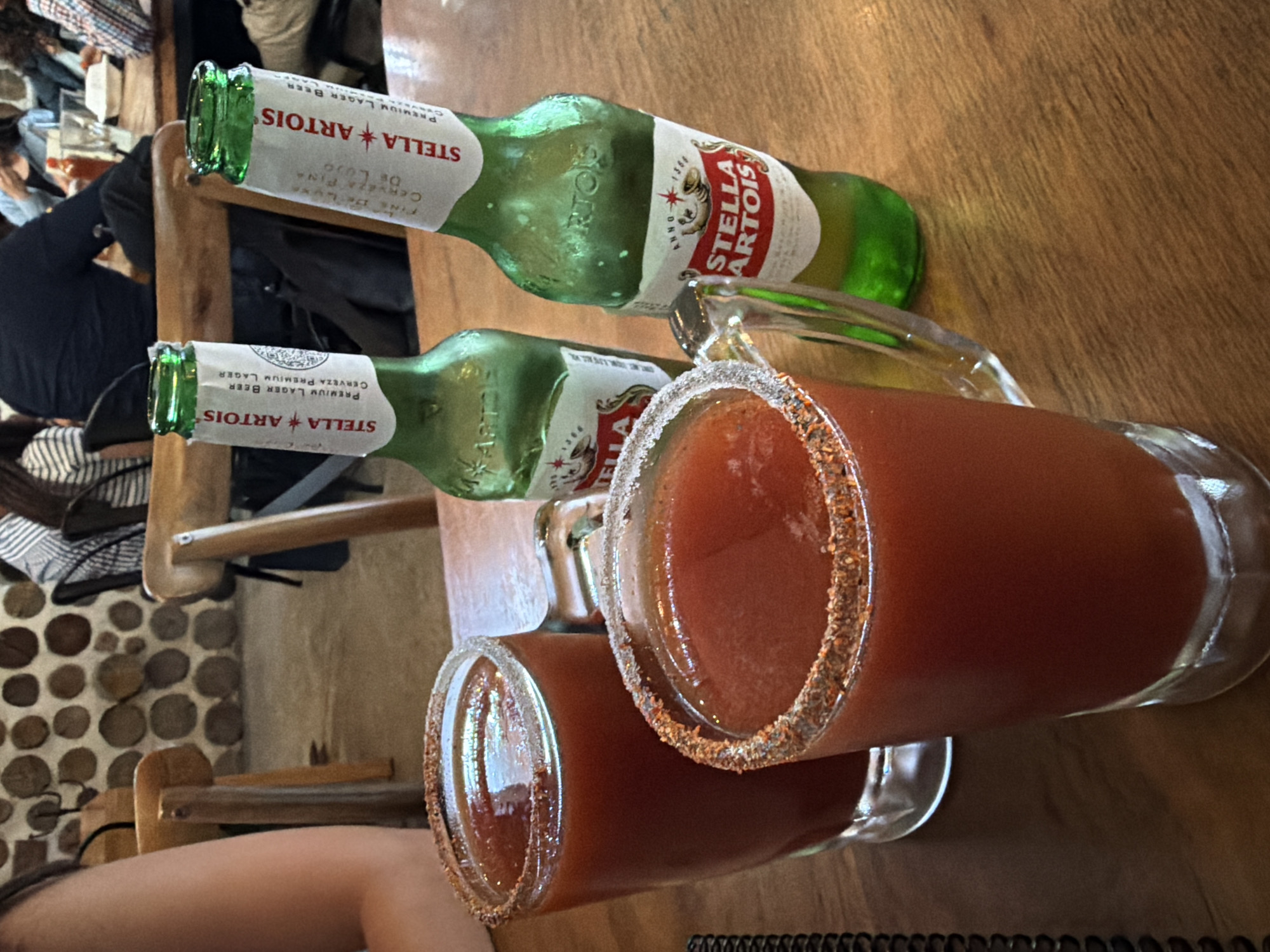 The image depicts a wooden table setting with two bottles of Stella Artois beer and two glasses filled with a red beverage, possibly Bloody Marys, garnished with a seasoned rim. The bottles are green with the Stella Artois label clearly visible. One glass is partially filled, while the other appears nearly full. In the background, there are glimpses of people, a wall adorned with circular patterns, and chairs. The scene appears to be in a restaurant or bar.