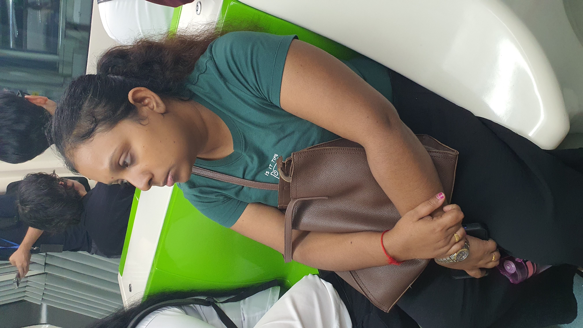 A medium shot captures a woman with dark hair pulled back in a ponytail, wearing a green t-shirt with white text that reads "IS IT TIME", as she sits on a bright green seat in what appears to be a public transport carriage. She is holding a brown leather handbag in her lap, with her arms crossed. A red thread is tied around her left wrist, and she wears a watch and two rings on her left hand. A pink water bottle is visible between her legs. The woman is looking to her right, her expression neutral. In the background, other passengers are visible, along with the train's interior features. The scene is likely within the Kuala Lumpur Mass Rapid Transit (MRT) system, given the distinct green and white seating design commonly found in its stations and trains, a key feature of the city's urban transit infrastructure.
