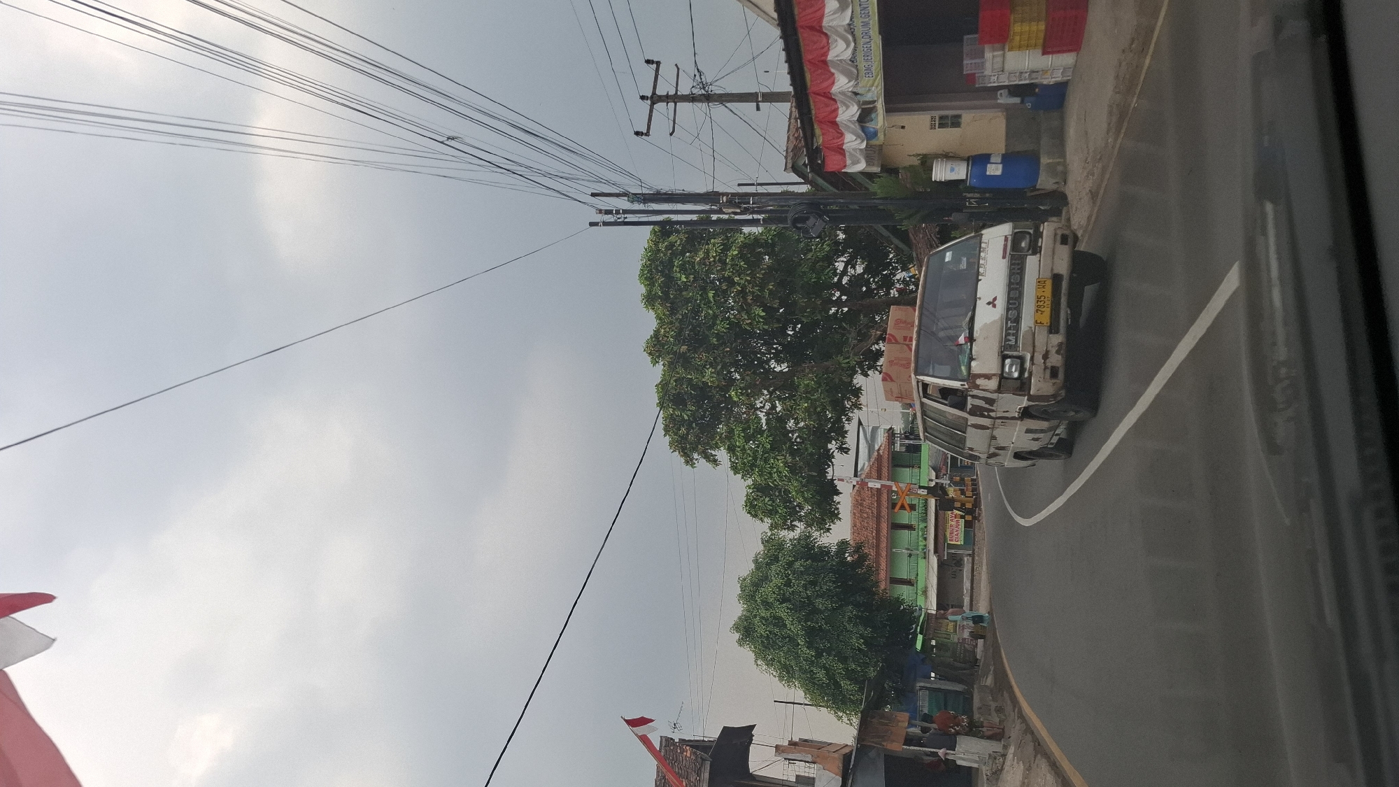 The image depicts a street scene in Cilaku, Indonesia, under an overcast sky. A white Mitsubishi van, showing signs of wear and rust, is driving on a paved road. The van has a license plate that reads "F 7935 WA". Several power lines crisscross the sky, supported by utility poles.
On the left side of the road, a building with a red and white flag flying from its roof is visible. Further down, a green building with a red roof stands. Trees with lush green foliage are scattered throughout the scene. On the right side of the road, another building features red and white decorative awnings. Several stacked crates, some red and yellow, are present in front of this building. A blue barrel and a white bucket are also visible. The road has a white line marking its center. The perspective suggests the photo was taken from inside a vehicle.