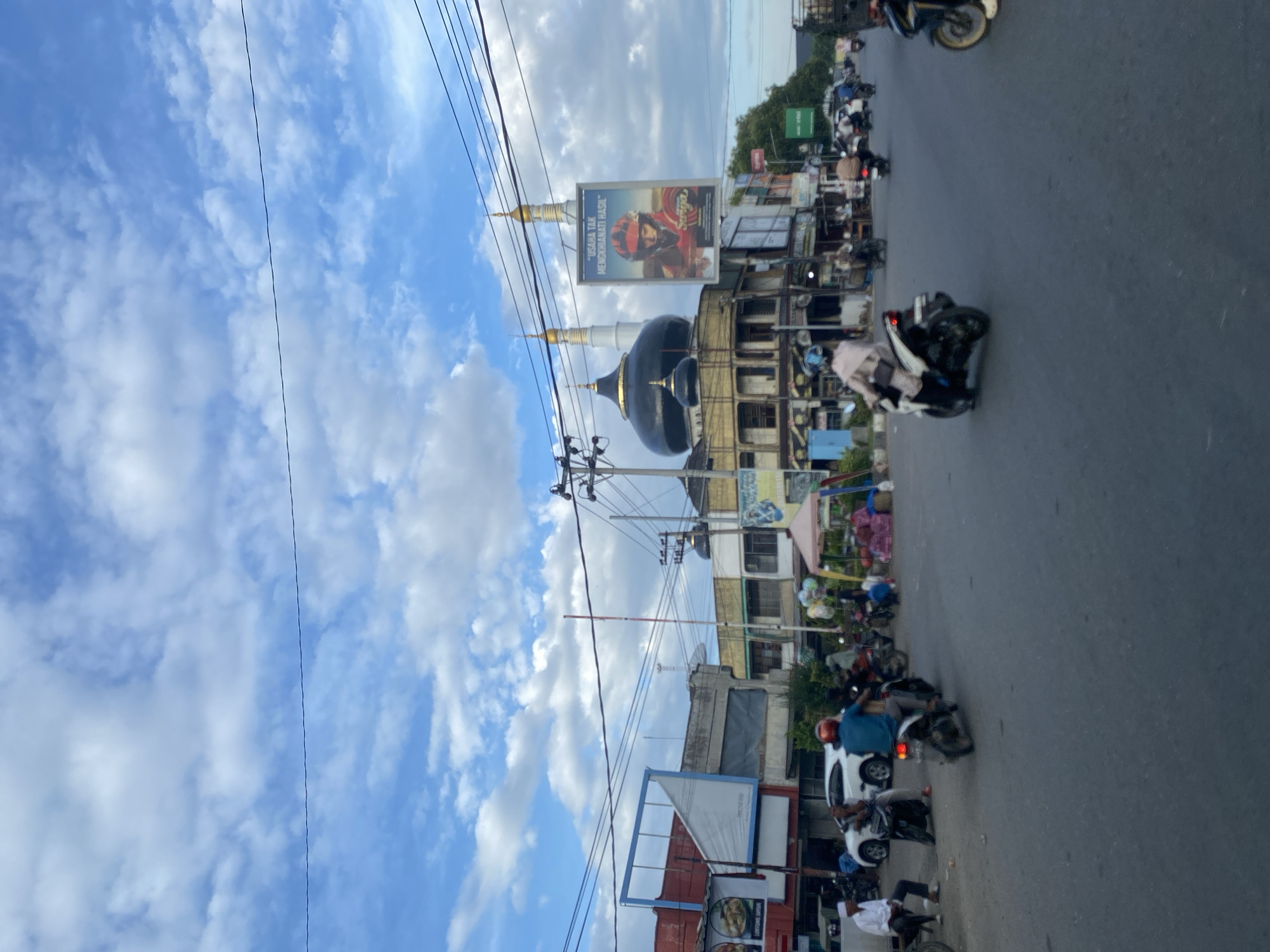 The image depicts a street scene in Pantegajah, Indonesia. In the foreground, there is a road with several motorcycles parked and moving. Buildings line the street, with shops and billboards visible. A black domed mosque with golden spires is visible in the mid-ground, with power lines running across the sky. The sky is cloudy.