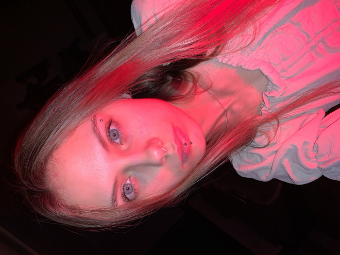 A close-up, angled portrait features a young adult female, located in the city of Foki, Russia. The subject has light skin, long brown hair, and blue eyes, which are prominently illuminated by a strong red light source positioned to the left. She is wearing a light-colored, possibly off-white, blouse with gathered fabric details visible on the upper chest and shoulders. Facial piercings include a silver hoop in the left nostril, two silver stud piercings on the left side of the upper lip, and two silver stud piercings on the right eyebrow. A thin silver chain necklace is visible around her neck. The subject maintains direct eye contact with the camera. The background is uniformly dark and indistinct, indicating a dimly lit interior environment.