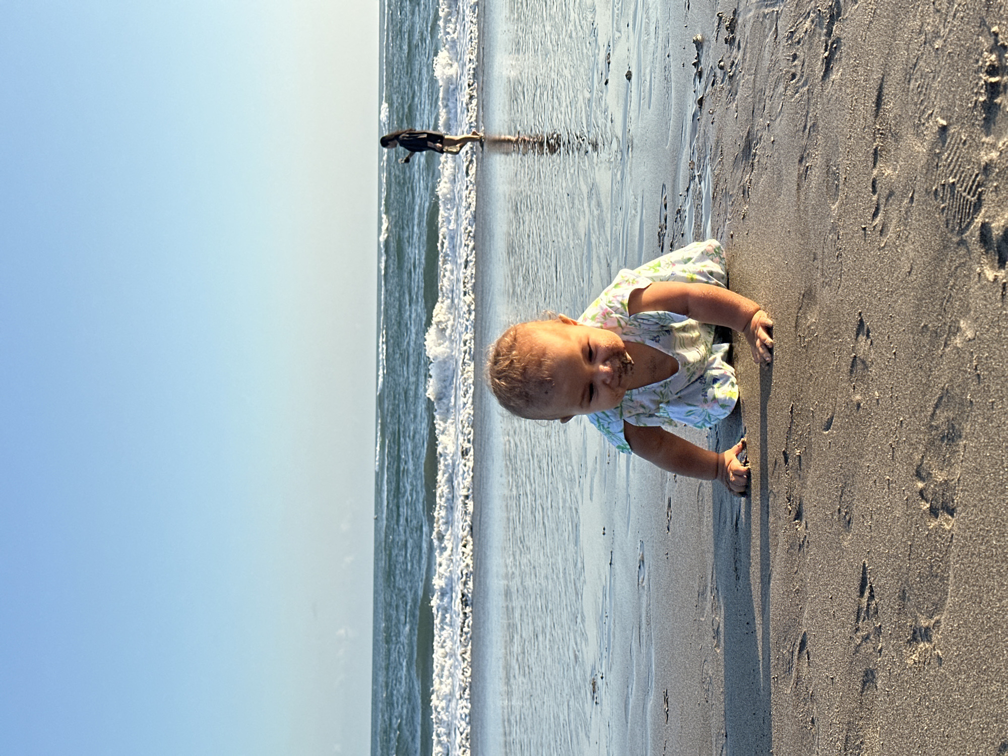 A baby crawls on the wet sand near the ocean on a beach in North Kuta, Indonesia. The baby is wearing a light-colored dress with a floral pattern and has sand on its face. In the background, a woman stands in the shallow water, looking out at the ocean. The waves are breaking on the shore, and the sky is a clear blue. The image is taken from a low angle, emphasizing the baby's perspective.