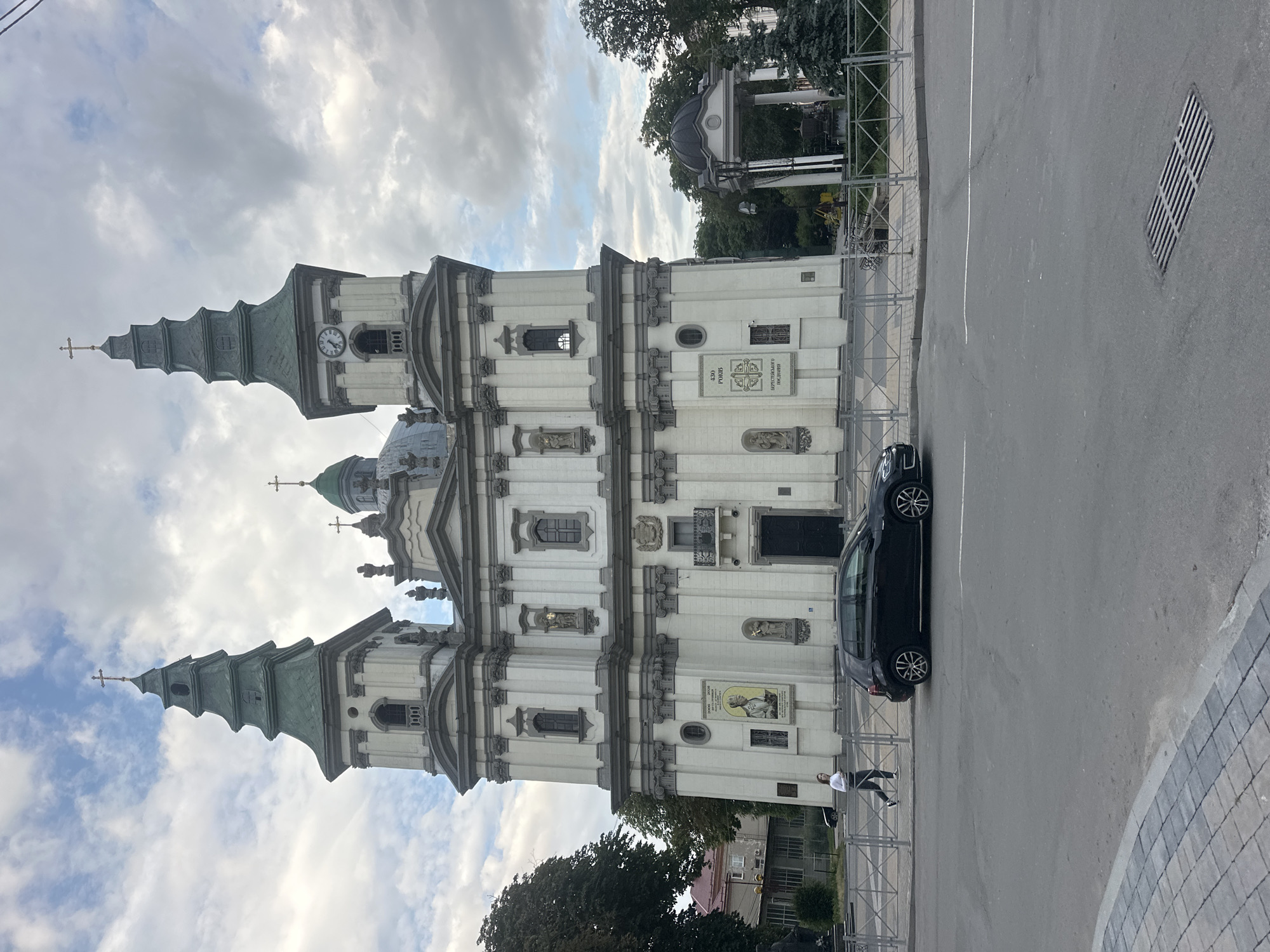 The image shows the Ternopil Cathedral in Ternopil, Ukraine, with its distinct Baroque architecture. Two prominent spires, topped with crosses, rise on either side of the central facade, one featuring a clock. The cathedral is painted white with gray architectural details. In front of the cathedral, a black car is parked on the street, and a woman is walking on the sidewalk. A metal fence separates the sidewalk from the street. In the background, trees and a small gazebo can be seen. The sky is overcast with scattered clouds.