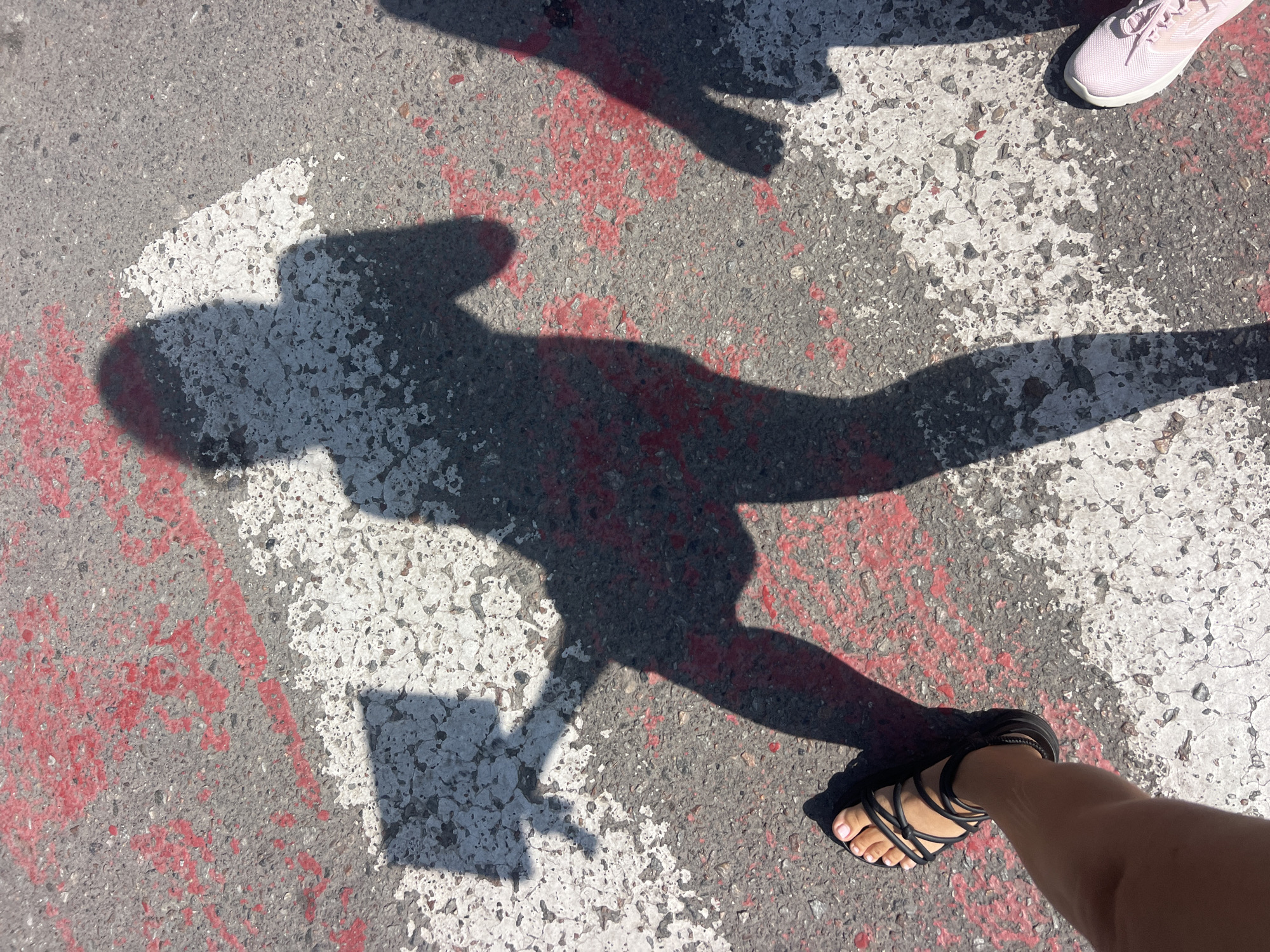 In Fontanka, Ukraine, a person's shadow is cast on asphalt marked with white and red paint, likely part of a crosswalk. The shadow depicts a figure holding what appears to be a phone or a similar flat object. To the left, a bare foot in a black sandal is visible, along with part of a leg. Another shadow, likely of a second person, is visible to the right, and a pink sneaker is also in the frame. The scene suggests a sunny day outdoors, possibly on a street or sidewalk.