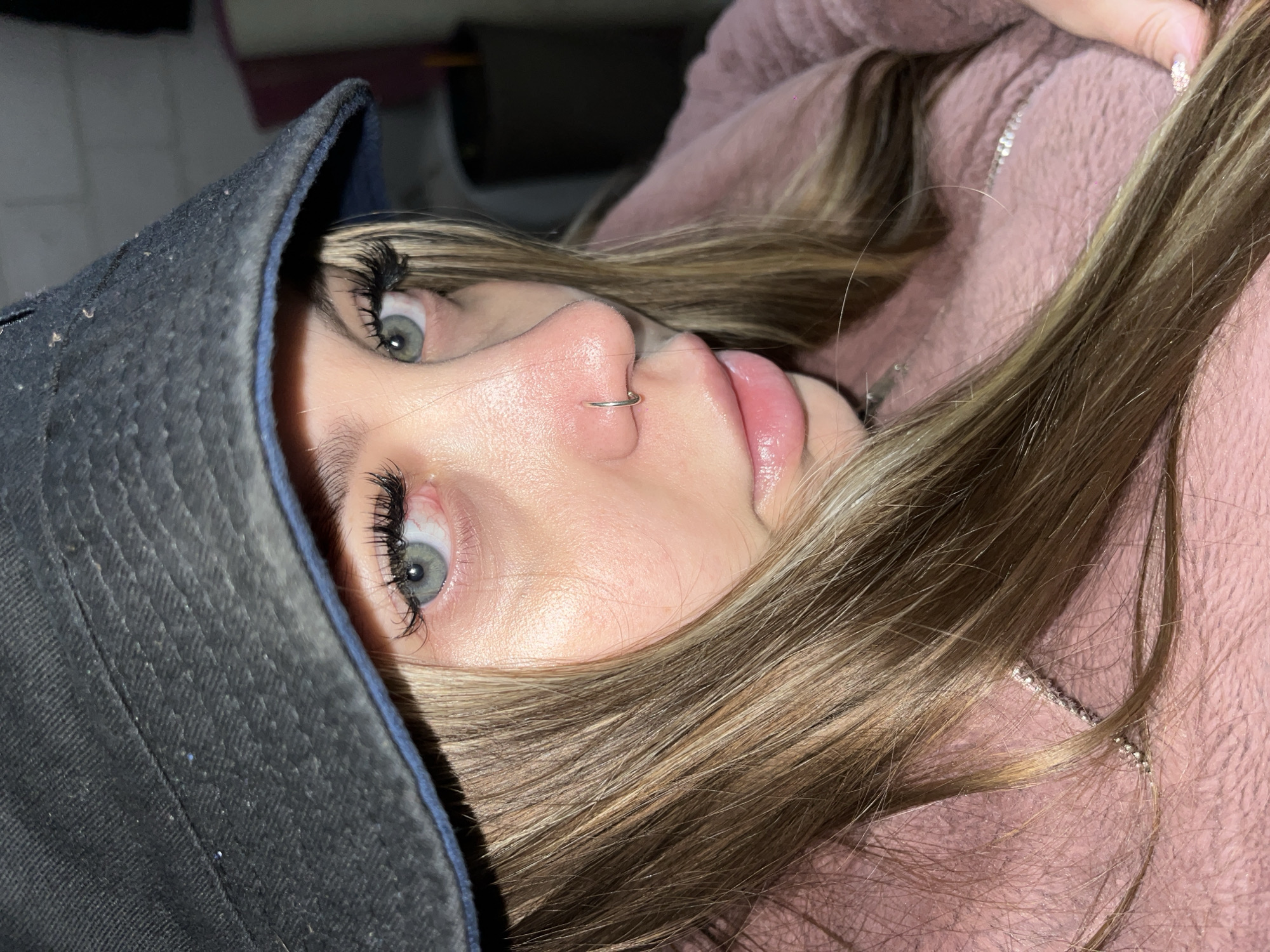 A young woman is captured in a close-up, slightly rotated perspective. She wears a dark-colored bucket hat that partially obscures her face. Her eyes are light, with full, dark eyelashes. A silver ring adorns her nose. Her lips are glossy and pink. She is wearing a pink, plush garment. Her hair is light brown, with lighter highlights. The background is partially visible, revealing indistinct shapes and lighting. The location is unknown and there is no evidence to suggest the location is Novopokrovskaya, Russia.