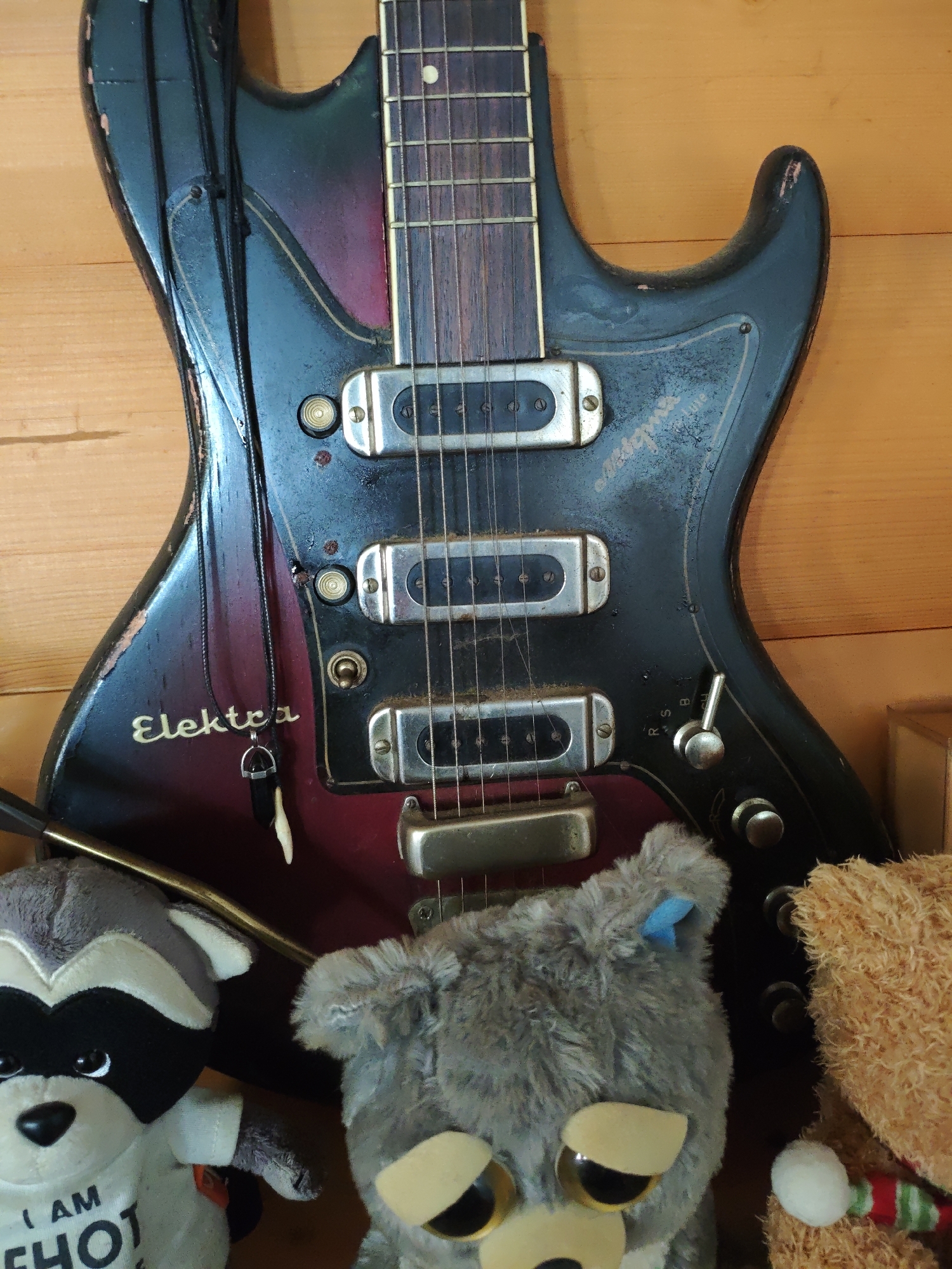 The image features an Elektra electric guitar positioned in front of a wooden wall. The guitar has a worn, vintage appearance, with a black and red body. Three silver-colored pickups are visible on the body of the guitar. The word "Elektra" is written on the body in yellow letters. A black necklace with a pendant hangs from the guitar. At the base of the guitar are three plush toys: a raccoon, an owl, and a bear. The bear has a red and green scarf.
The image does not provide any definitive information about the location being Боровково, Russia.