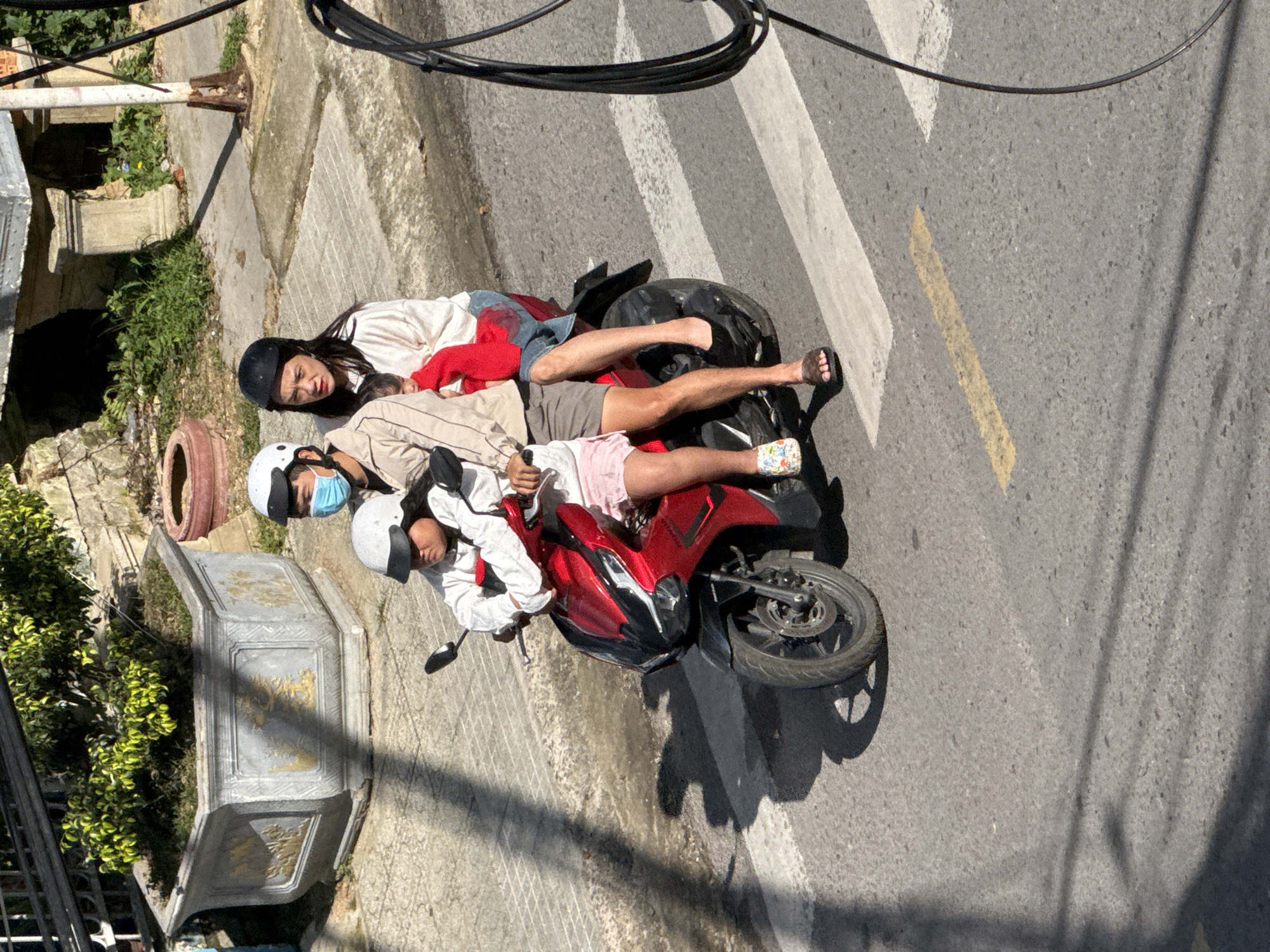 In Da Lat, Vietnam, three individuals, a driver, a passenger, and a child, are riding on a red scooter on a paved road with a pedestrian crossing. The driver, wearing a white helmet and a face mask, is controlling the scooter. The passenger, a woman with a black helmet, is seated behind the driver and holding a child wrapped in a red cloth. The child is also wearing a white helmet. The scooter is positioned on a crosswalk with white stripes. In the background, there are concrete planters with greenery, a stone wall, and electrical wires. The scene is illuminated by sunlight, casting shadows on the road.