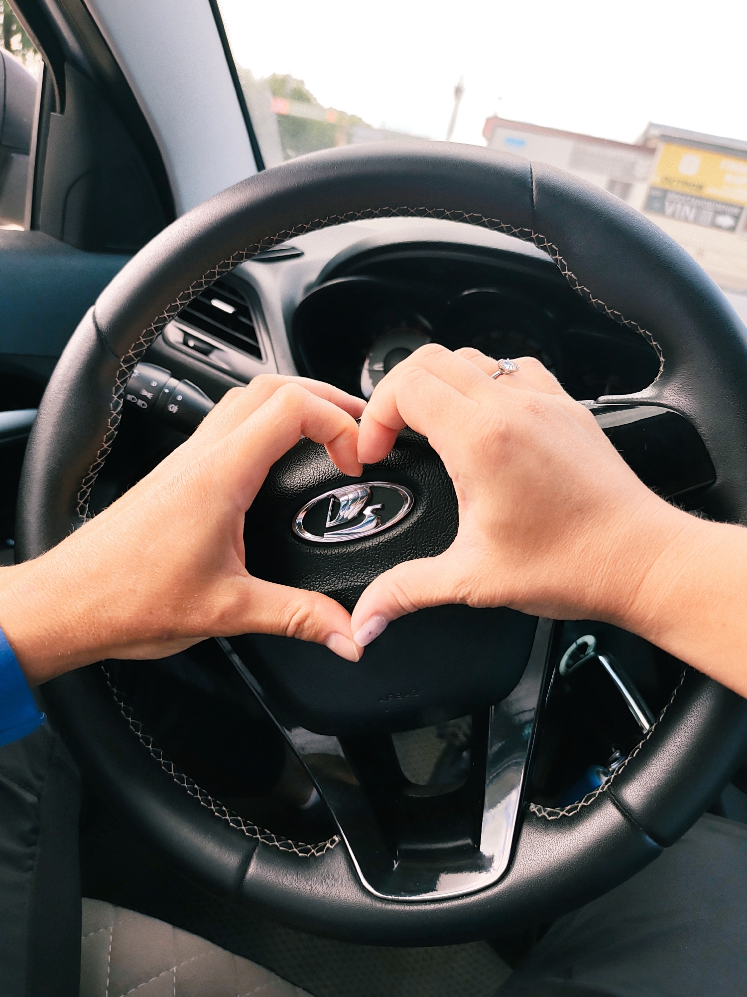 The image shows a close-up view from inside a car, focused on the steering wheel. Two hands are positioned to form a heart shape around the car's logo, which appears to be a stylized "LADA" emblem. The hand on the right has a ring on one finger. The interior of the car is mostly black, with some silver accents on the steering wheel. Outside the windshield, a blurred view of a building is visible. Based on metadata, the location is identified as the city of Chaikovsky, Russia. The subjects are the hands forming a heart shape on the steering wheel, and the implied subject is the car itself. The action is the formation of the heart shape. There is an interaction between the hands and the car's steering wheel. The environment is the interior of a car, with a blurred urban exterior visible in the background.