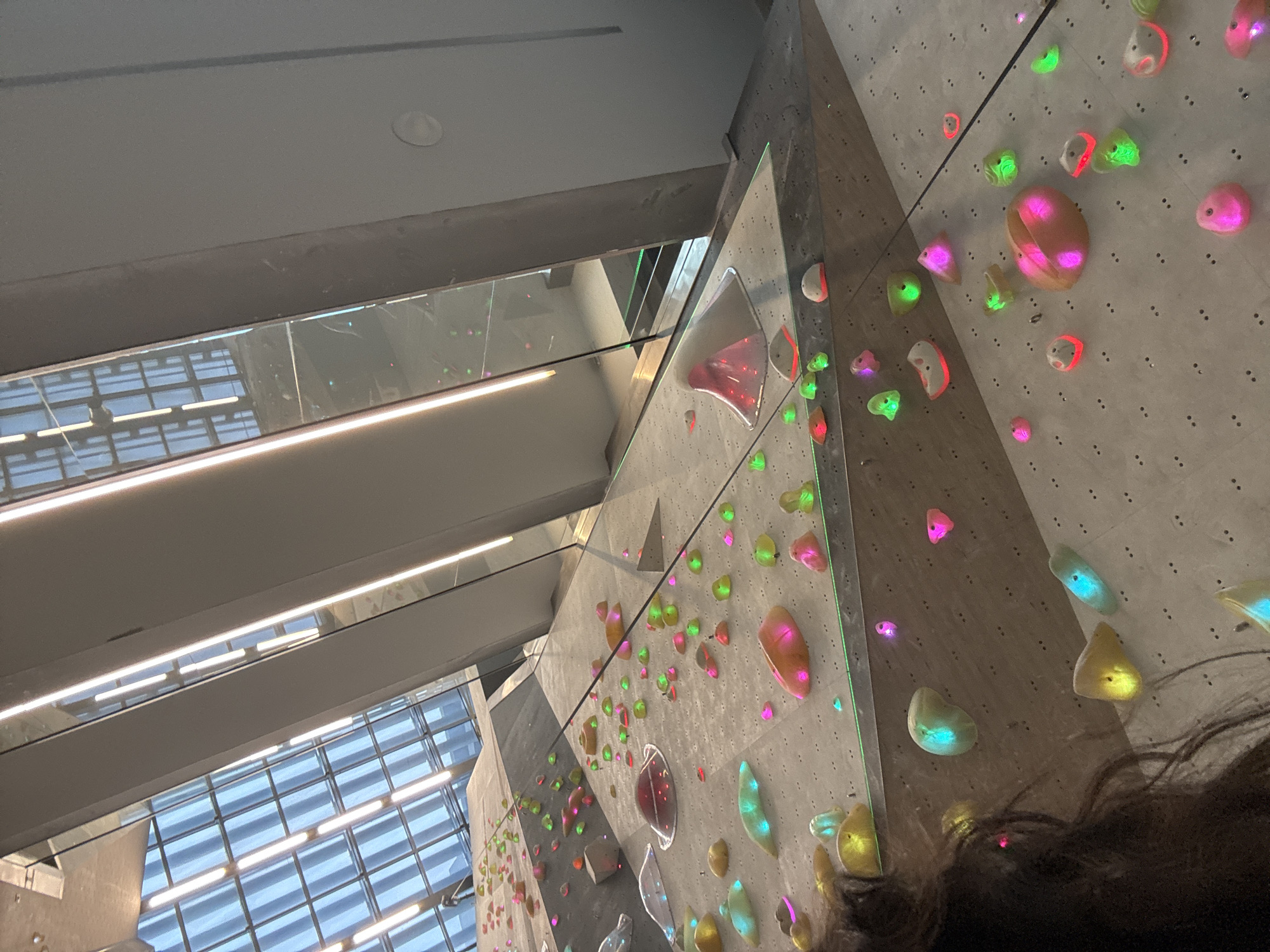 The image shows the interior of a climbing gym in Sofia, Bulgaria. The dominant features are the climbing walls, which are textured and adorned with various brightly colored climbing holds. The walls are angled, creating different degrees of difficulty. The background shows a glass-enclosed area with rows of lights and structural beams, suggesting a modern or industrial-style architecture. Some dark hair is visible in the lower right corner. The overall lighting appears to be a mix of natural light coming through the windows and artificial light from the ceiling. There are no discernible people, actions, or interactions directly visible in the frame, focusing on the structure and arrangement of the climbing facility.