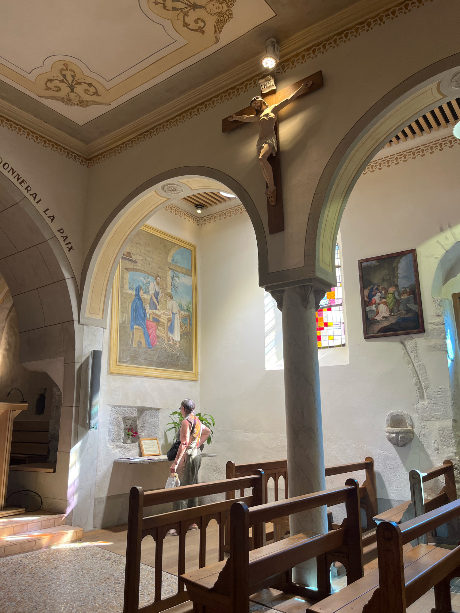 The image depicts the interior of a small, stone church in Yvoire, France, likely St. Pancras' Church given the architectural style. Sunlight streams in from a stained-glass window, casting colorful patterns on the floor and illuminating the space. A wooden crucifix hangs prominently on the wall above an arched opening. To the left, a large framed painting of the Holy Family is displayed. A woman, viewed from behind, stands near a niche containing a small floral arrangement and a framed certificate. She is wearing a patterned top and olive-colored pants and holds a white plastic bag. Wooden pews fill the foreground of the church. The walls and ceiling are adorned with painted decorative motifs. The overall atmosphere is one of peaceful reverence and historical charm.