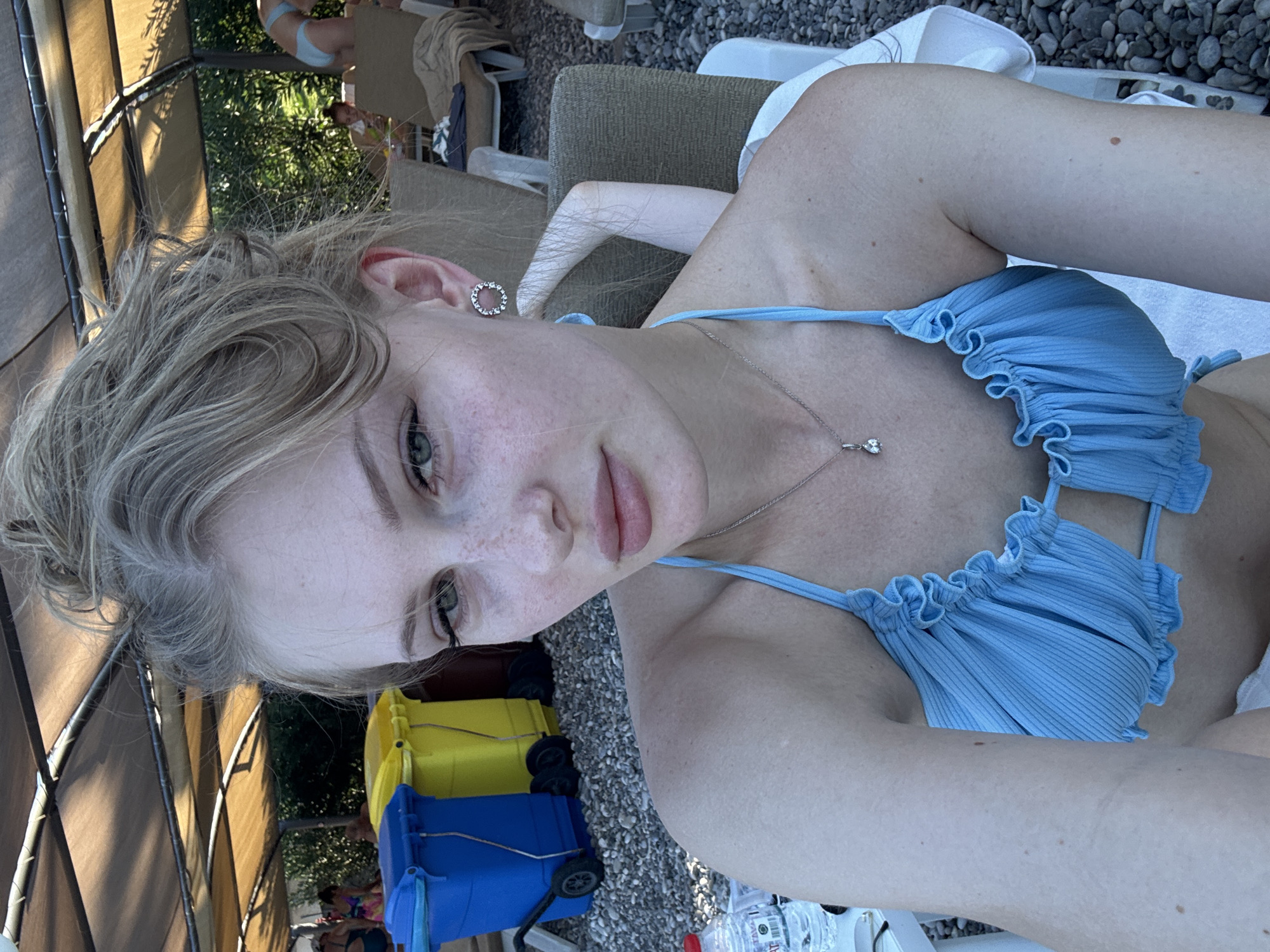 A young woman with blonde hair pulled up in a bun, wearing a light blue bikini top with ruffled trim, takes a selfie at an outdoor venue. She has light skin with freckles, green eyes, and is wearing a silver pendant necklace and hoop earrings. In the background, there are lounge chairs, some with towels on them, and a covered area with a brown canopy. To the left of the frame are two large trash bins, one blue and one yellow, on a pebble-covered ground. In the further background, other beachgoers can be seen, with one woman in a blue bikini and another in a colorful swimsuit. The environment suggests a beach or poolside location, likely in Kemer, Turkey, given the typical resort atmosphere and architecture.