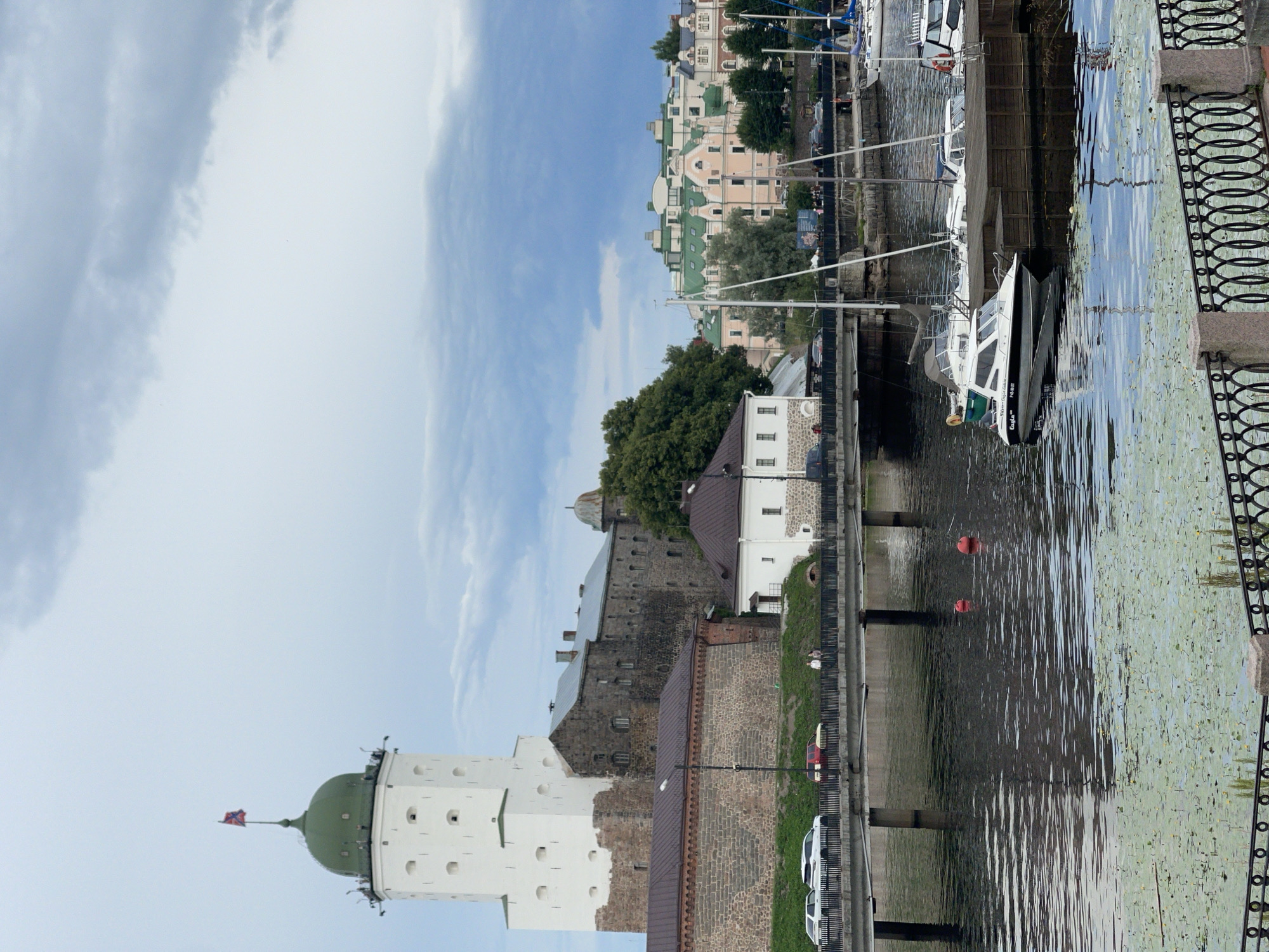 The image shows a view of Vyborg Castle in Vyborg, Russia. The castle is a prominent white tower with a green dome, flying a flag. Adjacent to the tower are stone buildings of the castle complex, some with dark roofs. A bridge with cars and pedestrians crosses a body of water in the foreground. Several boats, including sailboats and a smaller motorboat, are docked in the water. The water is partially covered with lily pads. In the background, there are buildings with light-colored facades and green roofs. The sky is overcast with clouds.