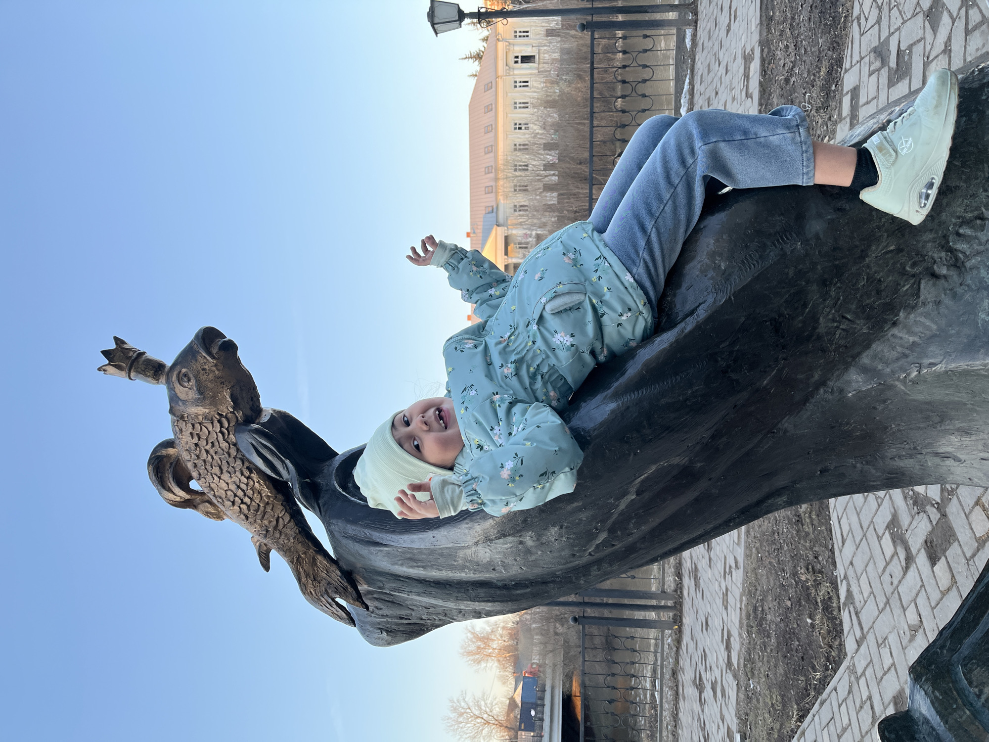 A young girl lies on a sculpture of a fish with a crown in Podolsk, Russia. The girl has a light blue jacket with a floral pattern and light blue jeans with light blue sneakers. The girl's eyes are open and she is smiling, raising her hand up. The fish sculpture is situated on a paved area near a metal railing and a body of water. A building and streetlamp can be seen in the background. The sky is blue.