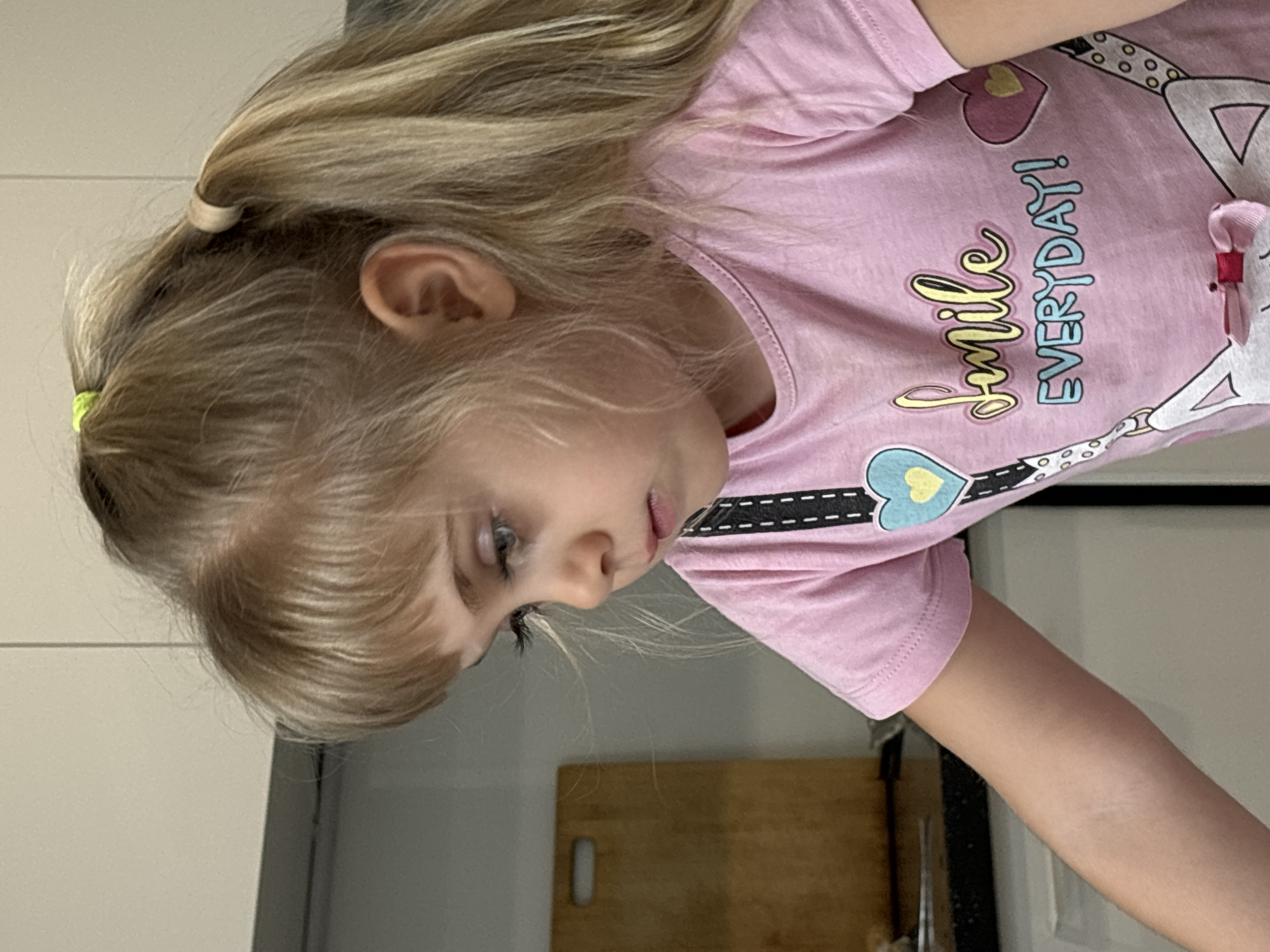 A young girl with blonde hair, partially tied back in a ponytail, is looking down. She is wearing a pink t-shirt with a cartoon design that includes the words "Smile Everyday!" The location appears to be indoors, possibly a kitchen, given the countertop and wooden cutting board visible in the background. No specific city can be identified.