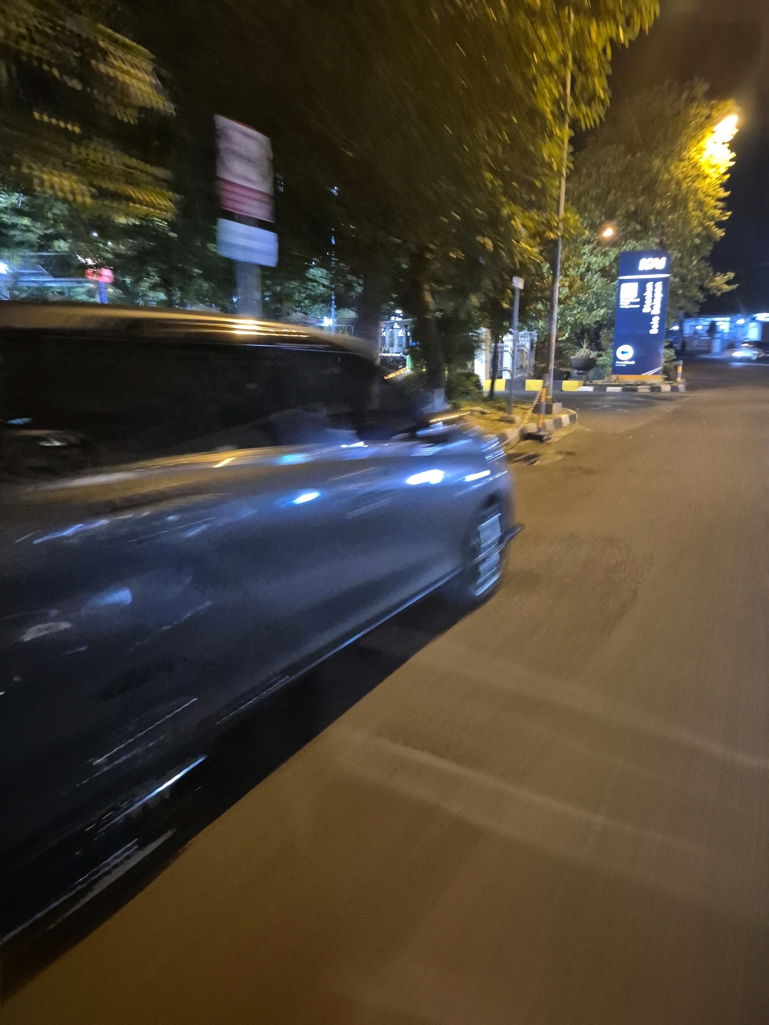 The image captures a night scene on a street in Surakarta, Indonesia. A dark-colored vehicle, possibly a sedan, is prominently featured, blurred due to motion. The vehicle's side is visible, reflecting the ambient lights of streetlights and the surrounding environment. The road surface is asphalt, marked with painted lines that are illuminated by artificial light sources.
To the right of the road, a tall, dark blue sign with white text stands at the entrance to an area. The text on the sign is partially legible and appears to indicate a location, possibly a public facility, with the word "Stasiun" visible, suggesting a train station. Below "Stasiun," the text "Solo Balapan" is clearly readable, identifying the specific train station as Solo Balapan, a well-known landmark in Surakarta. A circular logo with a white symbol is also present on the sign.
Behind the sign, trees with dense foliage are visible, their leaves illuminated by yellow streetlights. The lighting creates a dappled effect. Further in the background, structures and more trees are visible, with some areas appearing to be illuminated by blue lights, possibly from buildings or more streetlights. The overall atmosphere is nocturnal, with a mixture of dark shadows and bright artificial illumination.