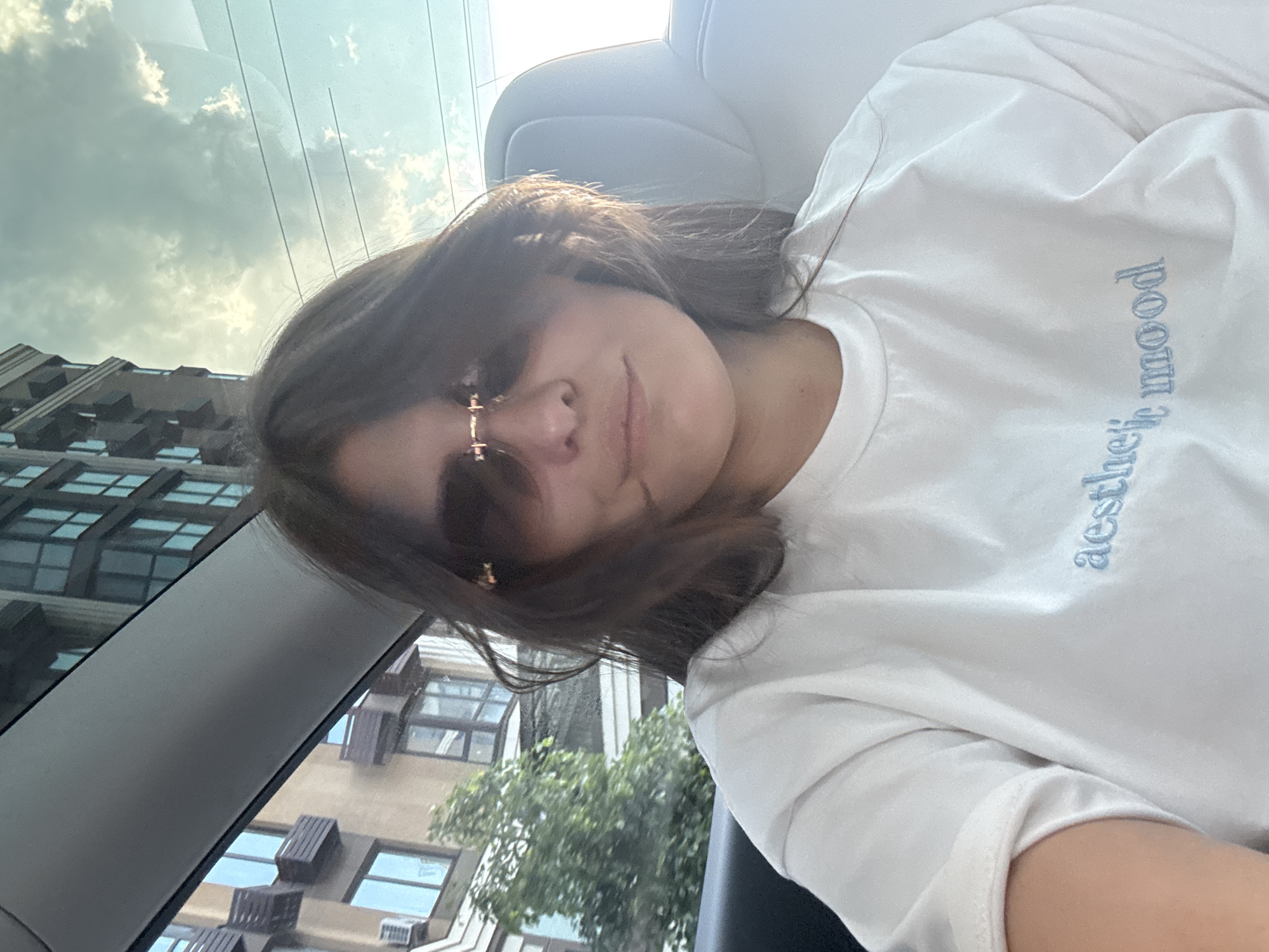 A young woman is captured in a selfie taken from inside a car, appearing relaxed with a slight smile. Her dark brown hair frames her face, and she is wearing fashionable, possibly frameless, sunglasses with gold-colored accents. She is dressed in a white long-sleeved t-shirt, which has the light blue embroidered text "aesthetic mood" visible on the chest.
The scene is set within a vehicle, with parts of the car's interior, such as a grey headrest and window frame, visible. Through the car window, a bright, cloudy sky dominates the upper background, indicating a daytime setting. Below the sky, urban buildings with multiple windows and some external air conditioning units are visible, along with green foliage from a tree, suggesting an urban or suburban environment. The overall atmosphere appears casual and contemplative, consistent with a relaxed moment in Sofiivska Borshchahivka, Ukraine.