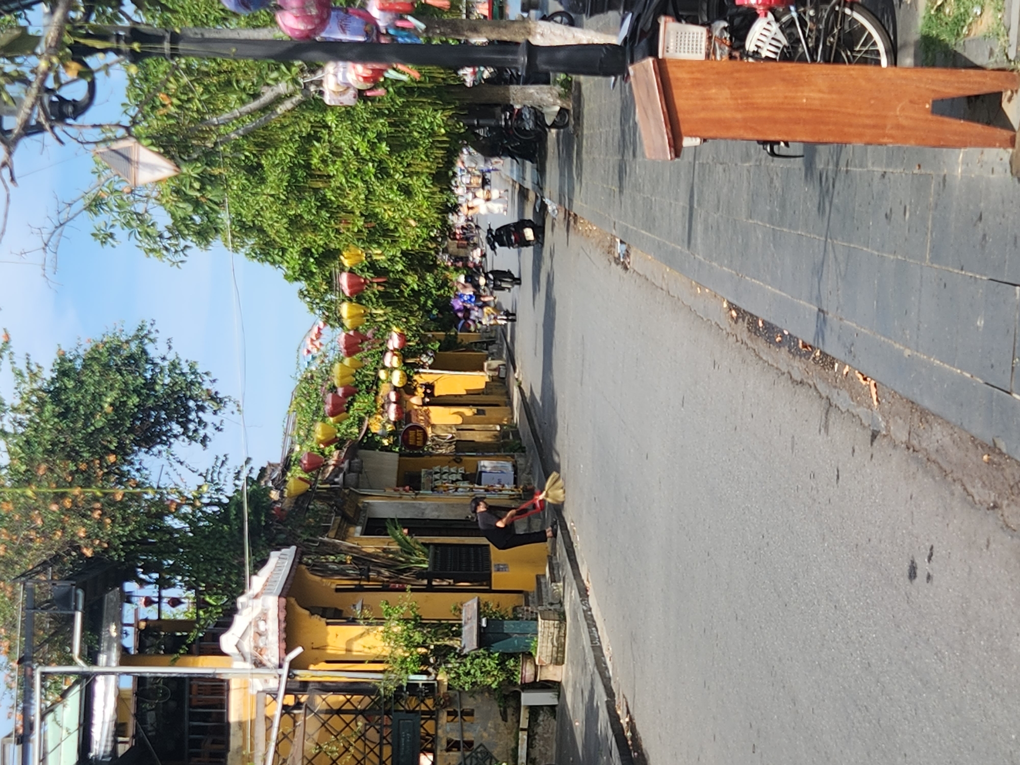 The image shows a street scene in Hội An Ward, Vietnam. A person dressed in black is sweeping the street with a broom. The street is paved with asphalt, and there is a sidewalk on the right side of the street. The buildings on the left are yellow and have ornate architecture, with balconies and lanterns hanging from trees. The street is lined with trees and lanterns, suggesting a festive atmosphere. Several motorbikes are parked on the sidewalk and in the street, and some are in motion. The sky is clear and blue.