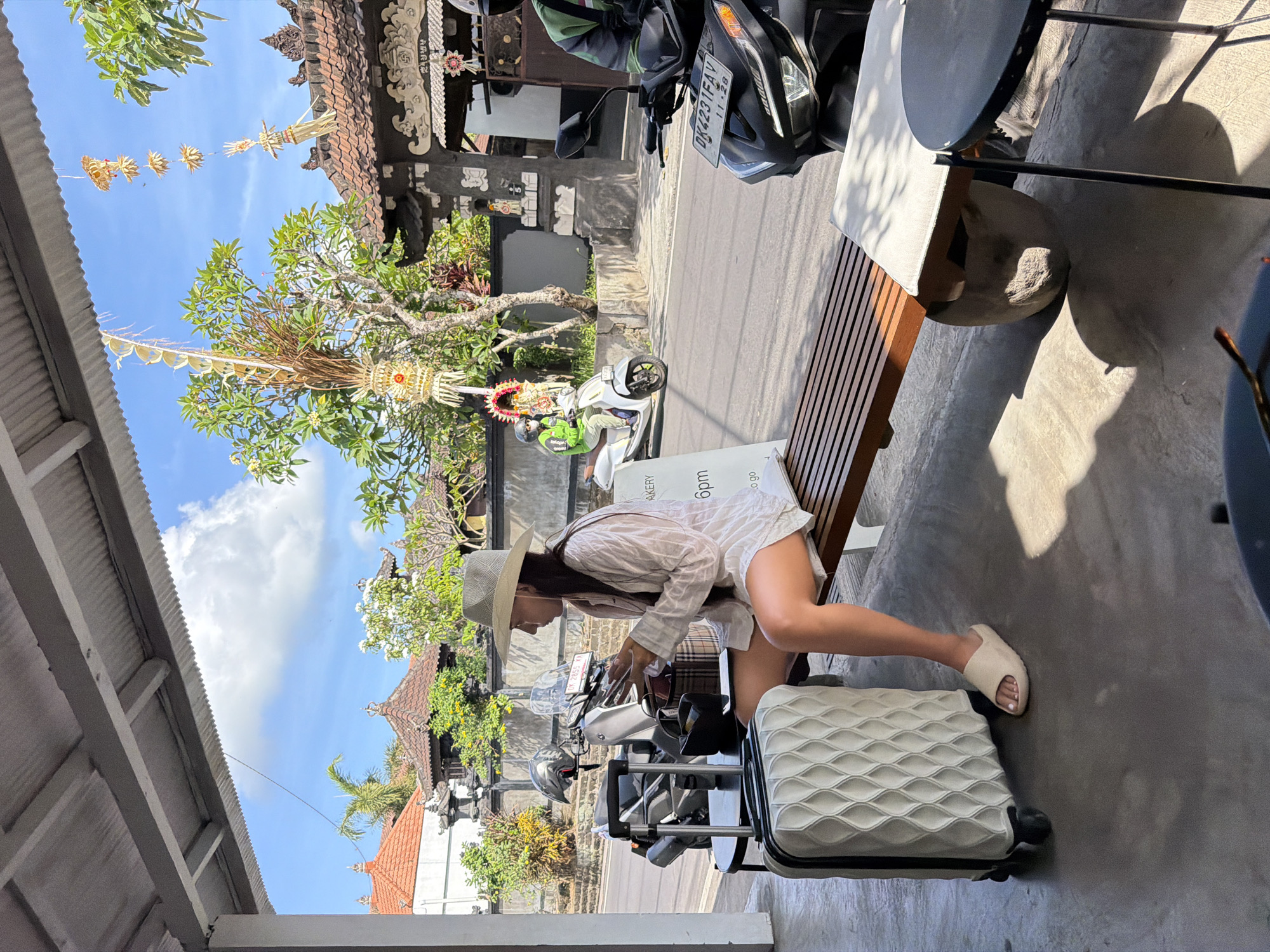In Mengwi, Indonesia, a woman sits on a wooden bench under a covered area, holding a phone and a wallet. A patterned white suitcase is beside her. In front of the bench is a black round table. The background shows a street with motorcycles, scooters, and traditional Balinese architecture including intricately carved stone gates and tiled roofs adorned with festive decorations. Lush green trees and a bright blue sky with scattered clouds complete the scene.
