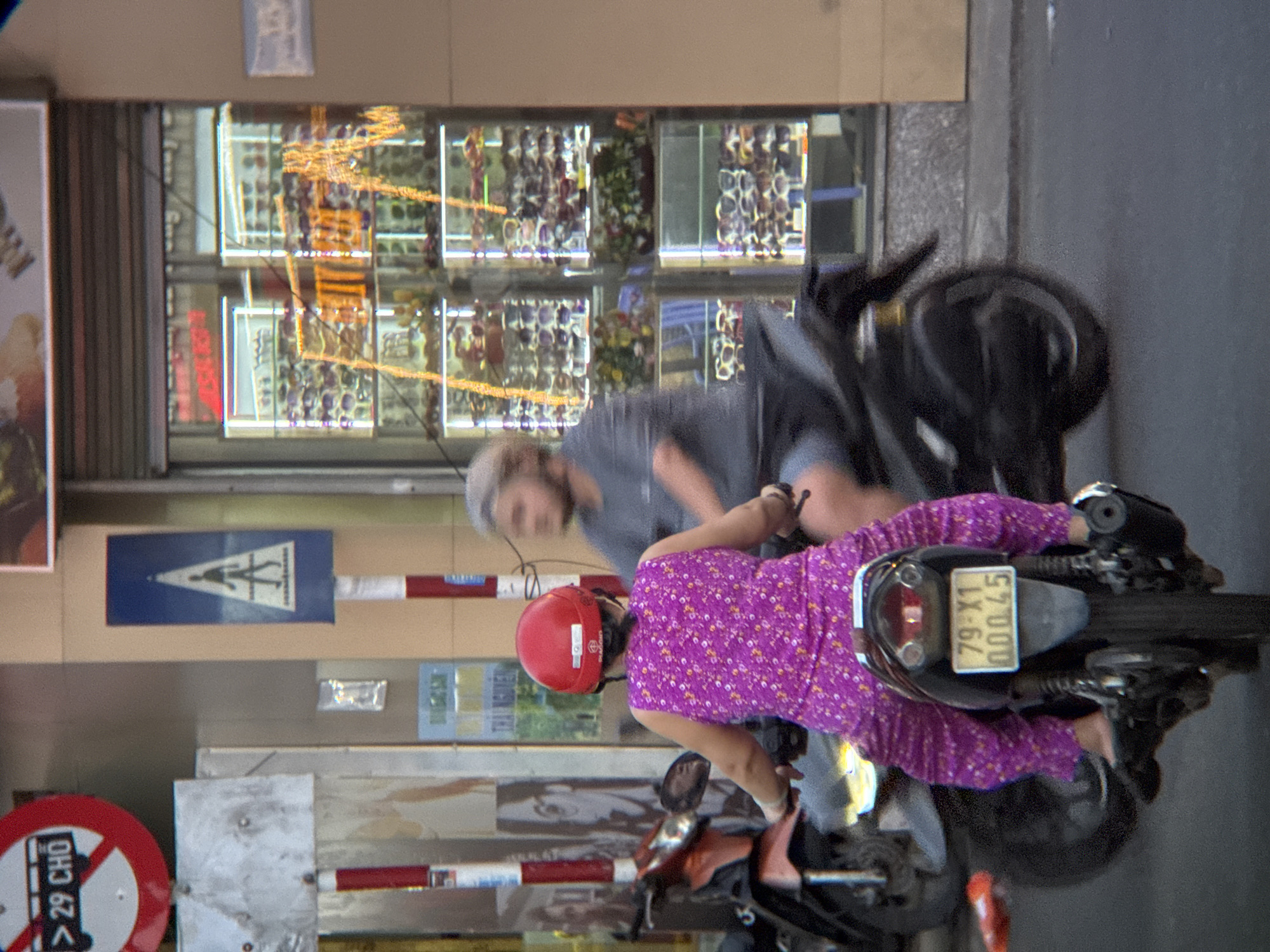 The image captures a street scene in Nha Trang, Vietnam, focusing on two individuals on motorcycles. In the foreground, a person wearing a red helmet and a vibrant purple, patterned outfit is riding a motorcycle. They are facing away from the camera, and their legs are visible, clad in the same patterned fabric. The rear of their motorcycle is equipped with a license plate displaying "79-X1 000.45". Behind them, another person, a male with a graying beard and wearing a gray t-shirt and shorts, is also on a motorcycle and appears to be following or riding alongside.
The environment suggests an urban setting. To the left, a red and white striped pole stands near a sign with the text "> 29 CHỢ" on a white circle with a red border. Further back, a store window displays numerous pairs of sunglasses on shelves, illuminated by warm lighting, suggesting an optician or eyewear shop. The storefront itself has a dark, possibly metal, roller door partially open. A pedestrian crossing sign with a silhouette of a person walking is also visible on a blue background with a white striped pole. The ground is asphalt, indicating a road or street. The overall lighting suggests daytime.