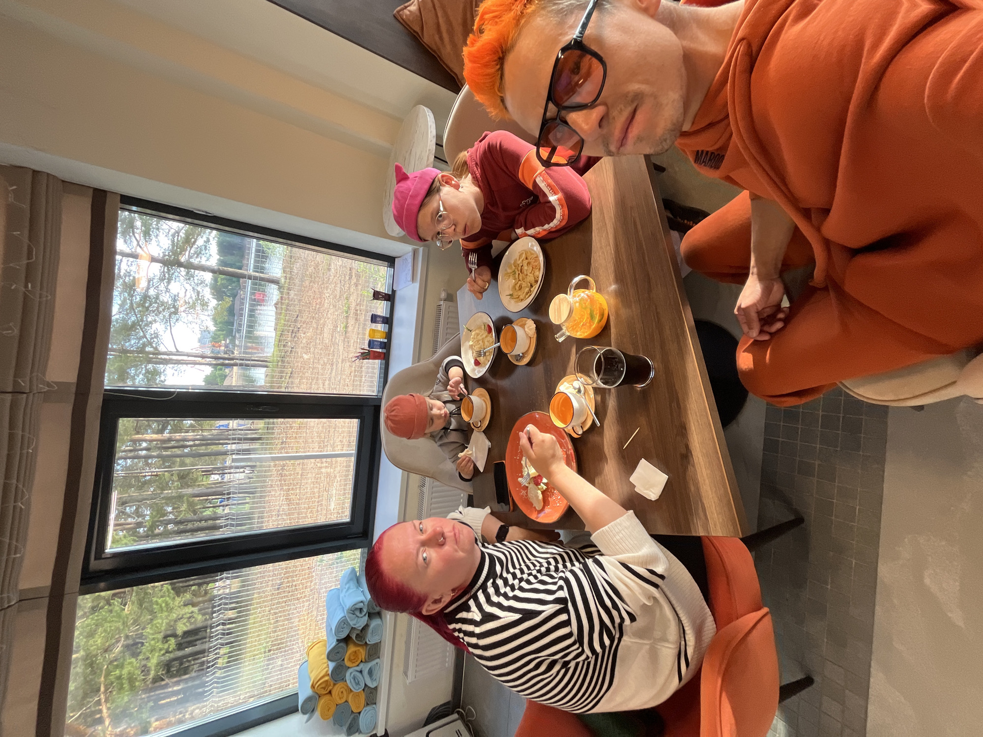 An overhead, slightly angled shot captures an indoor dining scene featuring four people of varying ages gathered around a wooden table, enjoying a meal or snack. In the immediate foreground, a man with bright orange hair, wearing sunglasses and an orange tracksuit with "MAROO" partially visible on it, looks directly at the camera.
Opposite him, a younger individual with a pink hat and glasses is focused on eating a pasta-like dish from a white plate. Further down the table, a woman with red hair and a black and white striped top also looks towards the camera while eating a dessert from an orange plate. A small child, wearing a red hat, is seated in a high chair at the table, also engaged with food and a cup.
The wooden table holds several plates of food, teacups (some orange, some white), a glass teapot with a wooden lid, another ceramic teapot, and glasses. Napkins and a toothpick are also present.
A large window dominates the left side of the frame, revealing an outdoor scene with numerous trees and what appears to be a fence or structure beyond. Horizontal blinds are visible on the window panes. Below the window, neatly rolled towels in blue, yellow, and white are stacked on a shelf. The room is brightly lit by natural light, suggesting it is daytime. The floor visible near the table features small, grey square tiles. The overall impression is a relaxed, familial gathering in a modern setting.