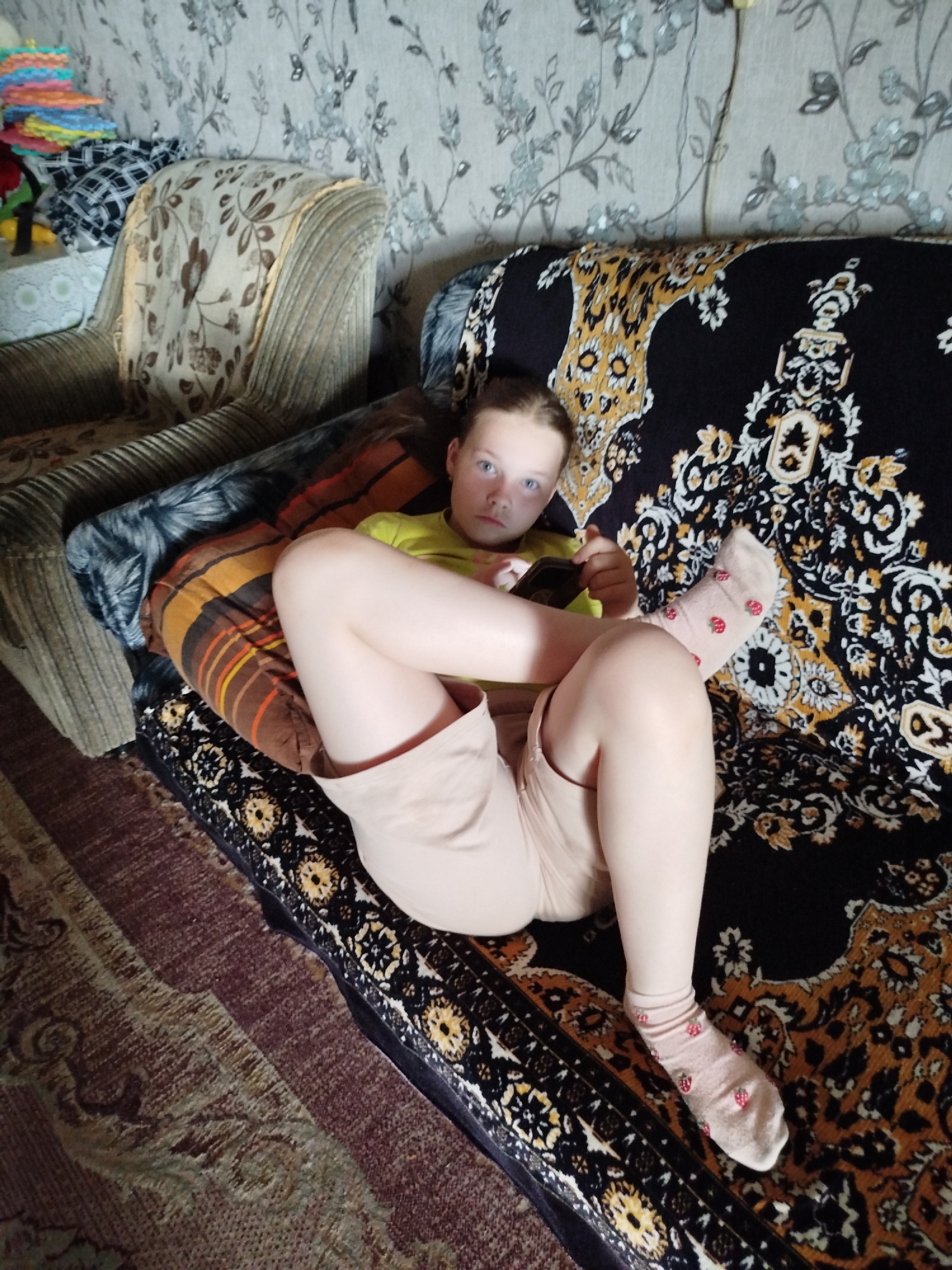 A young girl is lying on a couch in a room. She is wearing a yellow t-shirt and light-colored shorts. She is holding a phone in her hands and looking at it. The couch is covered with a black and gold patterned blanket. There is a striped pillow under her head. The room has floral wallpaper and a patterned carpet on the floor. A chair with a floral pattern is visible to the left of the couch. The scene appears to be indoors in a residential setting.