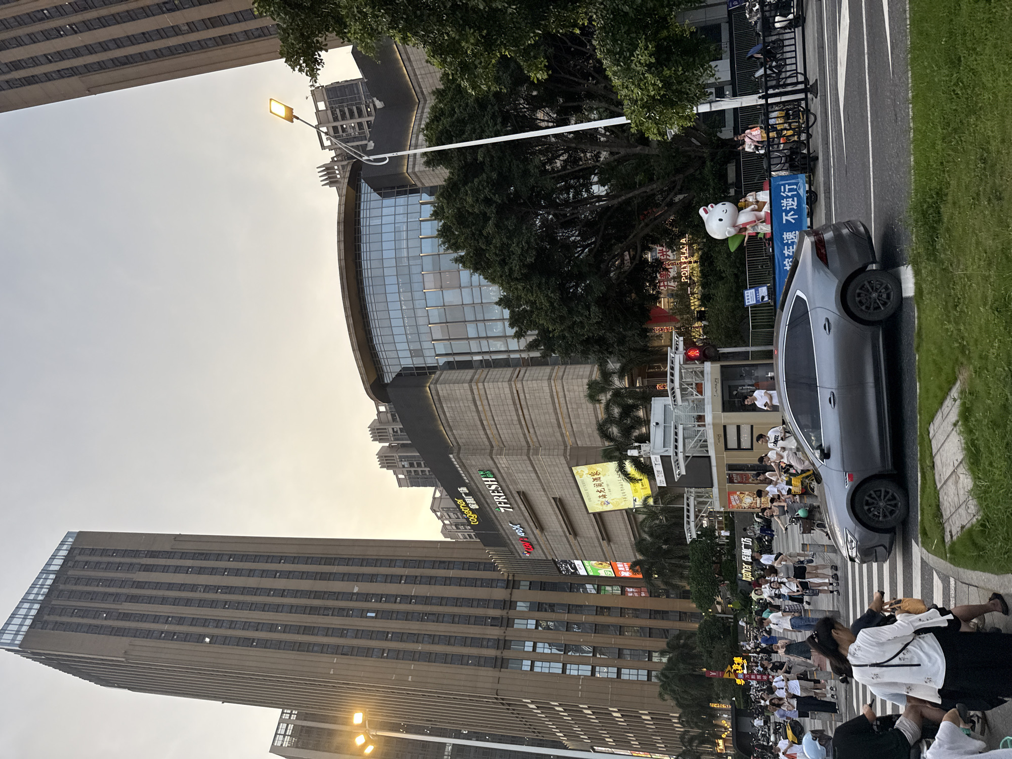 Here is a description of the image:
The image depicts a busy street scene in the Haizhu District of Guangzhou, China. Tall buildings, including the "Poly Plaza" building complex and other structures, frame the sides of the photo. A crowd of people and scooters are gathered along the sidewalk. A gray car is parked on the side of the street, close to an advertisement display for the location. Street signs and commercial signage are also visible. The time of day appears to be late afternoon or early evening, as indicated by the lighting.
