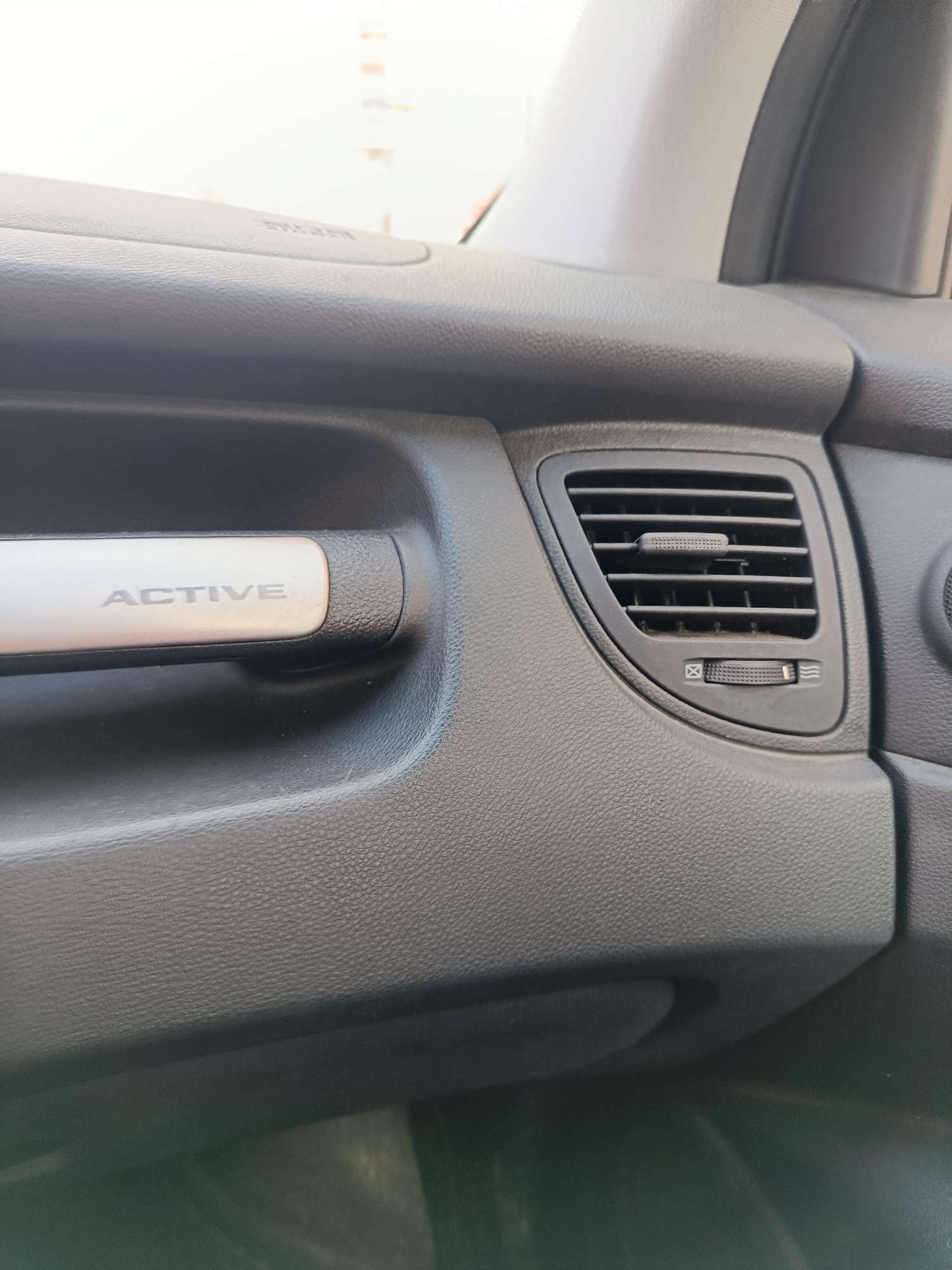 This media file is a close-up, eye-level photograph of the interior of a car, specifically focusing on a section of the dashboard. The primary subjects are car interior components. We see a textured dark gray plastic dashboard, a silver trim piece with the word "ACTIVE" inscribed on it in a light gray font, and a car air vent with adjustable louvers and a circular dial control below it. The dial has symbols that likely indicate airflow direction or temperature.
The scene is static, with no activity or event taking place. The lighting appears to be natural daylight, suggesting it's daytime. There are no visible people, animals, or explicit weather cues. The location context of Orekhovo-Zuyevo, Russia is noted but not visually evident within the frame itself. The overall impression is of a functional, everyday car interior. The textures of the plastic and the brushed metal trim are clearly visible. The focus is sharp on the "ACTIVE" trim and the air vent, with the rest of the dashboard softly blurred. There is no discernible emotion conveyed. The only visible text is the word "ACTIVE" on the silver trim.