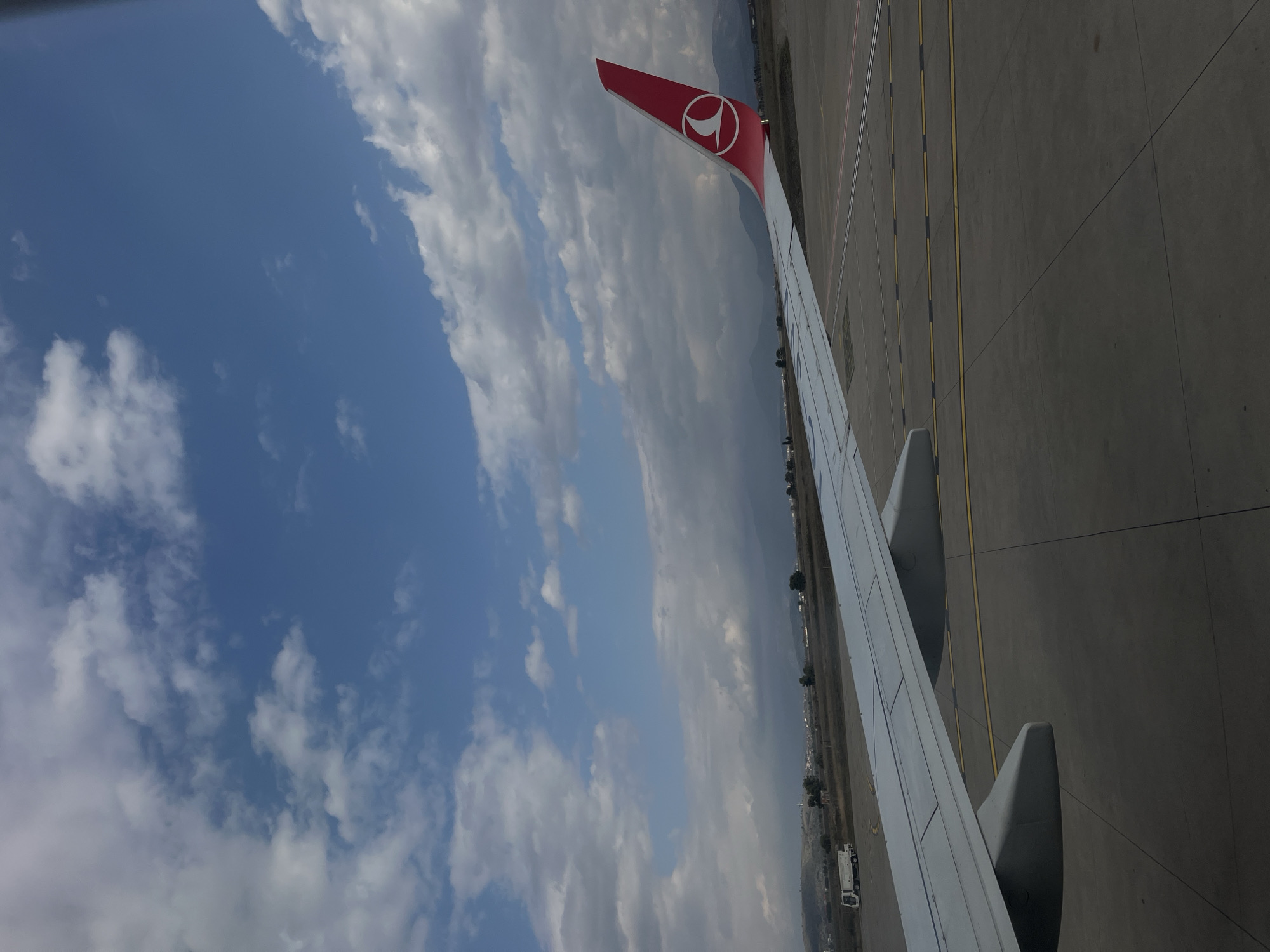 The image displays a view from an aircraft window at an airport in Donja Cijevna, Montenegro. The right wing of a passenger jet, light grey with two engine nacelles visible beneath, extends diagonally from the lower left to the center-right. The aircraft's vertical stabilizer (tail fin) is prominent on the right, colored red and featuring a white Turkish Airlines logo. The aircraft rests on a dark grey concrete or asphalt apron marked with multiple parallel yellow lines and some faded pink/white lines. In the distant left, a white utility vehicle is parked on the apron. The background beyond the airfield comprises a landscape of dry, sparse vegetation and scattered distant structures, including a white building with a pointed roof or spire on a prominent hill. Dark, undulating mountain ranges define the furthest horizon. The sky overhead is partly cloudy, with patches of clear blue interspersed with white and grey cumulus clouds. The aircraft appears stationary. No individuals are visible.