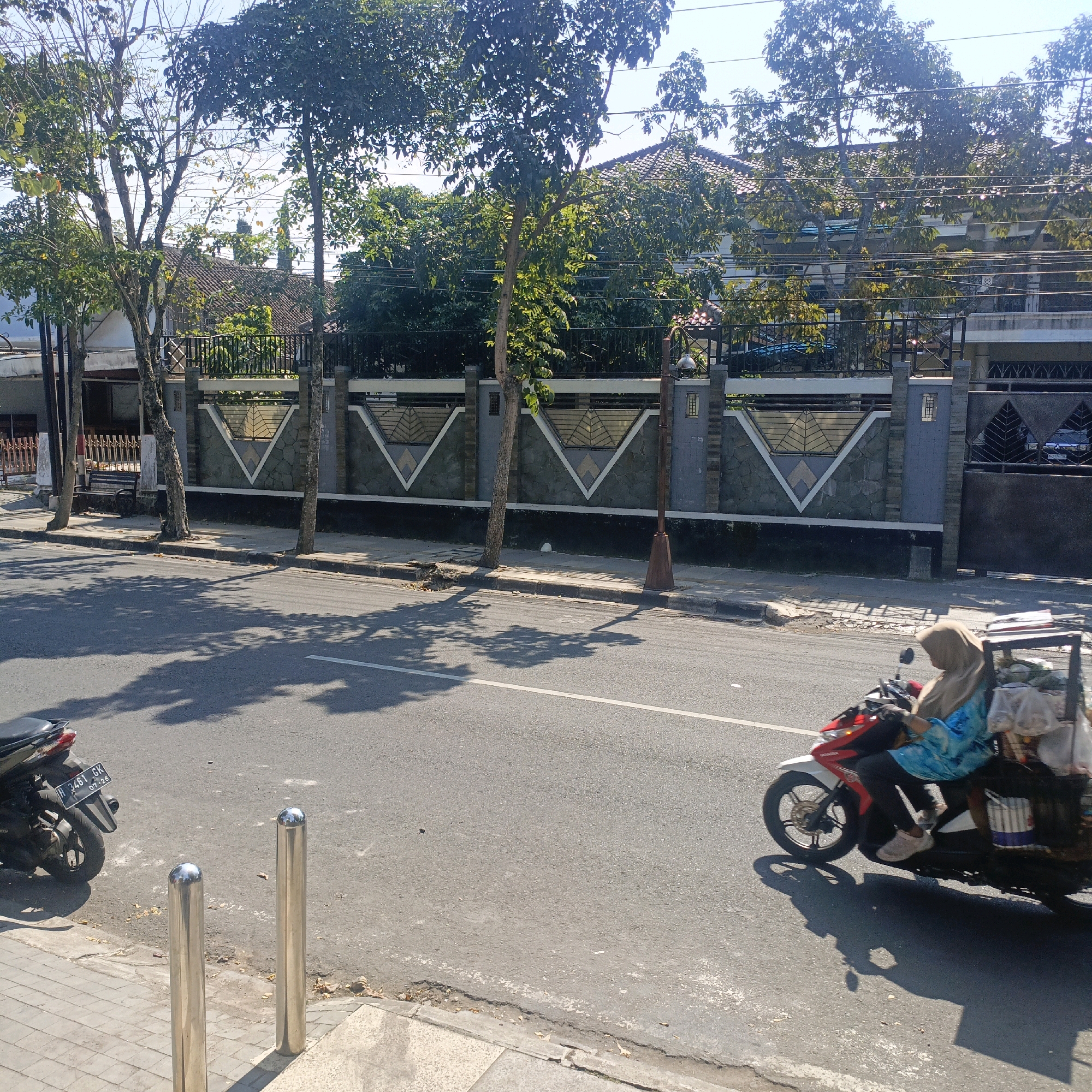 A street scene in Salatiga, Indonesia, is captured on a sunny day. A person wearing a hijab and a blue and white patterned top rides a red and white scooter. The scooter is fitted with a rack carrying bags of what appear to be groceries or merchandise. The scooter is in motion, casting a distinct shadow on the asphalt road. To the left, a parked motorcycle with the license plate "H 9461 GK" is visible. The road is lined with a sidewalk featuring two cylindrical metal bollards. Beyond the sidewalk, a decorative fence with geometric patterns separates the street from a residential area with buildings and lush green trees. The fence is constructed with stone and concrete pillars. The sky is clear and bright blue.
