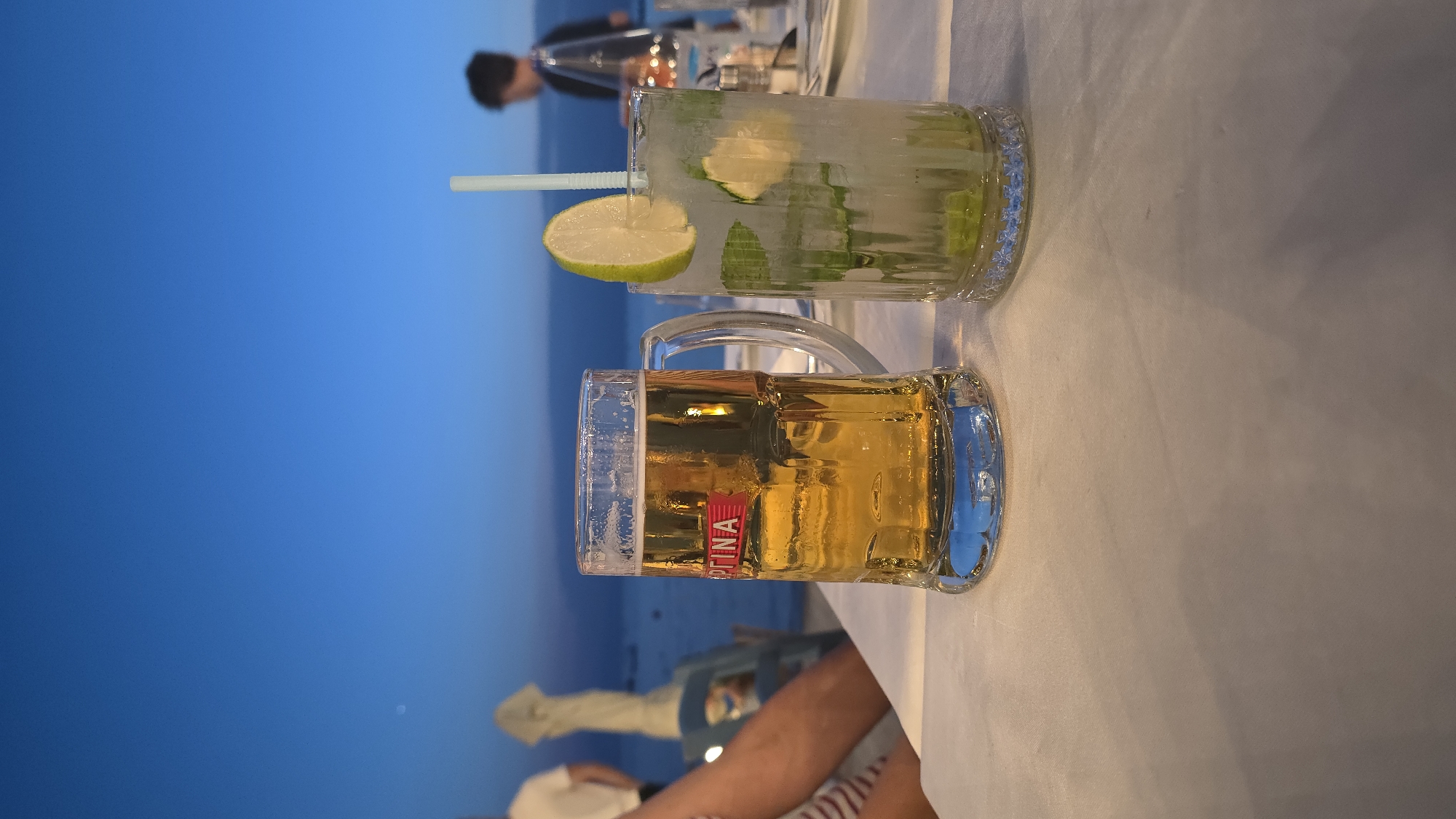 A high-angle, medium shot captures two drinks on a white tablecloth at dusk. To the left, a tall, clear glass mug filled with golden beer, a red label visible on the side. To the right, a tall glass of a light-colored cocktail, garnished with a lime slice and mint leaves, with a blue straw. The background reveals a calm, blue body of water, likely the Aegean Sea, with a faint horizon line. In the distance, silhouettes of people and beach umbrellas are visible. The sky is a deep blue, transitioning into darker tones at the top, suggesting the twilight hours. The scene evokes a relaxed, outdoor dining experience, possibly at a beachfront restaurant in Kassandra Municipal Unit, Greece.