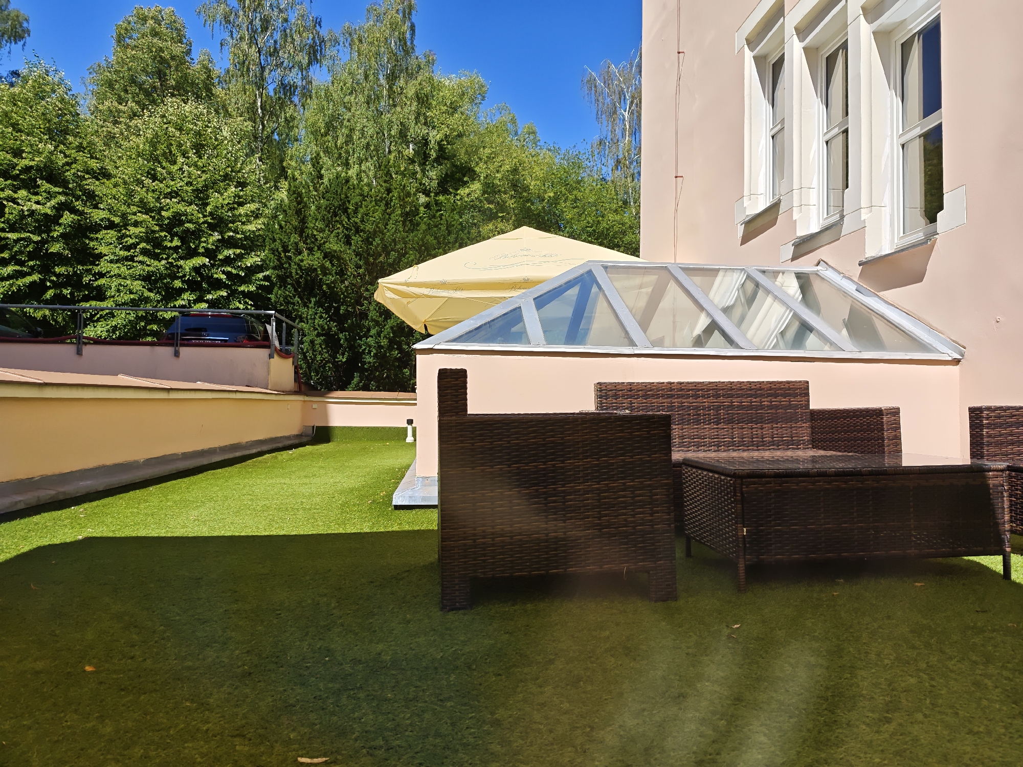 The image depicts an outdoor seating area with artificial grass in Karlovy Vary, Czechia. The area is furnished with dark brown wicker outdoor furniture, including a sofa, an armchair, and a coffee table. A glass-covered structure, possibly a conservatory or sunroom extension, is visible behind the furniture, attached to a pale pink building. The building has three tall windows with white decorative trim on the upper floor. A large yellow umbrella is partially visible behind the glass structure, suggesting it provides shade. In the background, a line of lush green trees and a clear blue sky dominate the scene. A railing and a portion of a car are visible to the left of the seating area, indicating a boundary or access point. The sunlight casts distinct shadows across the artificial grass.