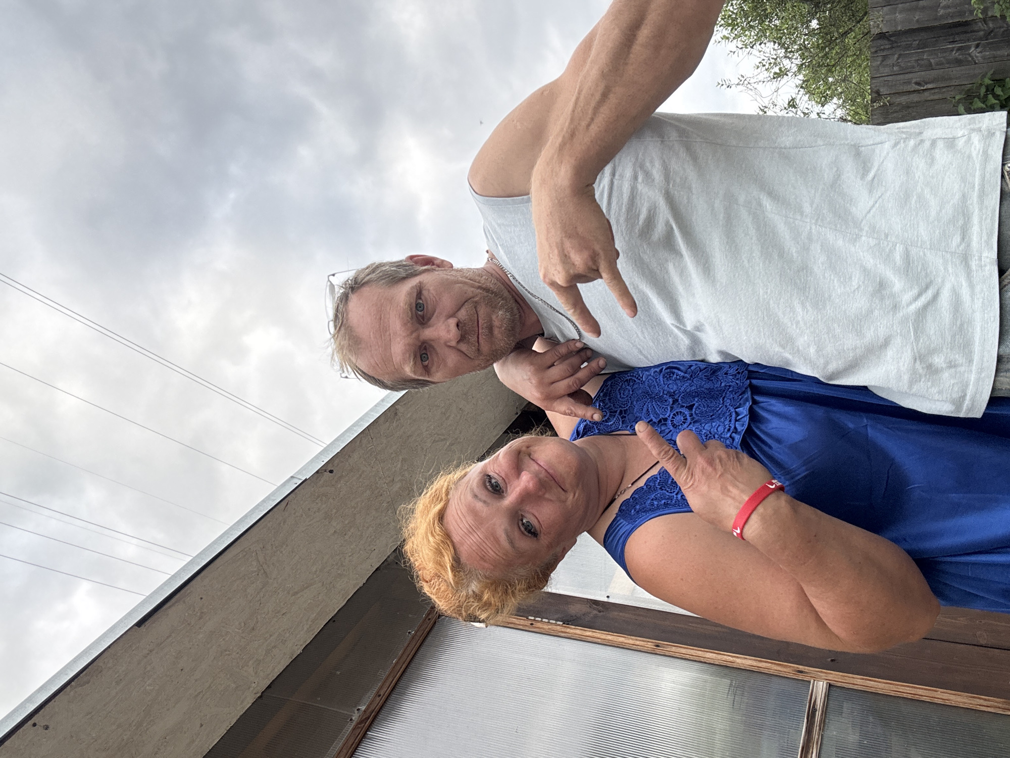 In Chaikovsky, Russia, a woman with bright red, permed hair and a blue lace-trimmed dress poses for a selfie with a man. She wears a red wristband. The man, with light brown hair and stubble, wears a light grey t-shirt and has glasses perched on his head. He makes a rock-on hand gesture. They are positioned in front of a corrugated plastic-covered structure, with a wooden frame and an overhanging roof. Power lines are visible against a cloudy sky. A wooden fence and green foliage are present in the background.