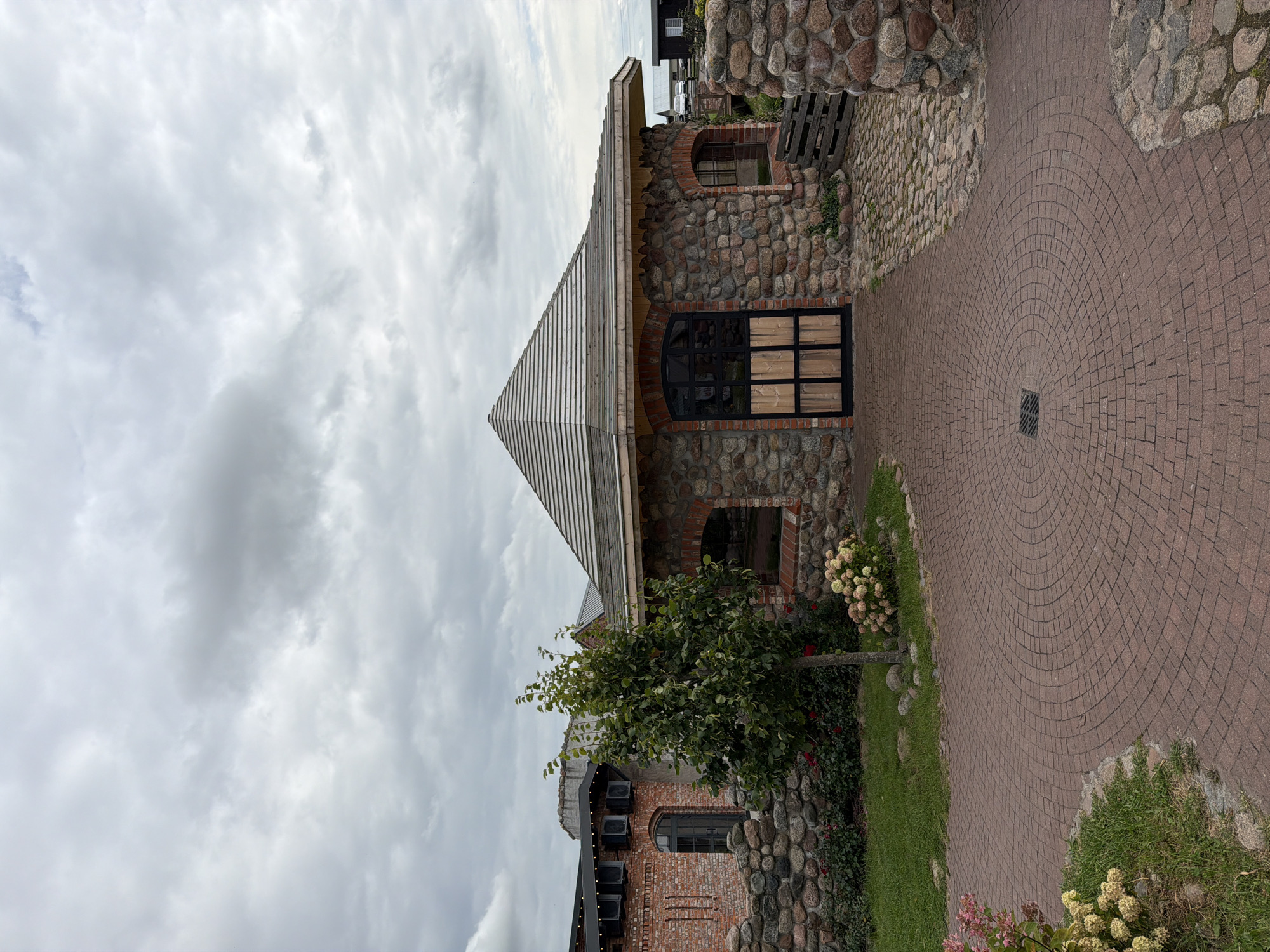 In Nekrasovo, Russia, a rustic stone building with a conical roof stands under a cloudy sky. The building features a wooden and glass door and arched windows. A circular brick path leads to the entrance, surrounded by grass and stone walls. A small tree and flowering bushes are visible on the left. The scene depicts a quiet, outdoor setting with traditional architecture.