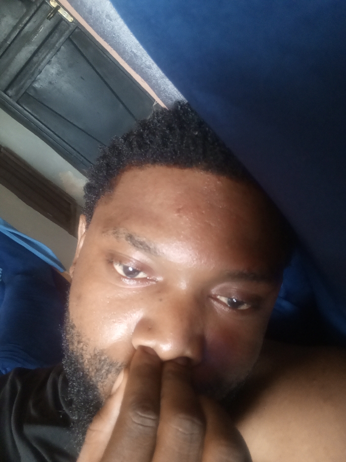 The image displays a close-up portrait of an adult male, taken in Magboro, Nigeria. The subject has dark skin, short, dark curly hair, and a beard. He is partially clad in a black garment. His right hand is positioned with fingers touching his nose and mouth area. His eyes are open, directed towards the viewer. The background on the upper left reveals a dark, horizontally paneled structure, possibly a wooden door, with a metallic latch visible. The upper right background is occupied by dark blue fabric. Illumination is uneven, with a brighter light source from the left.