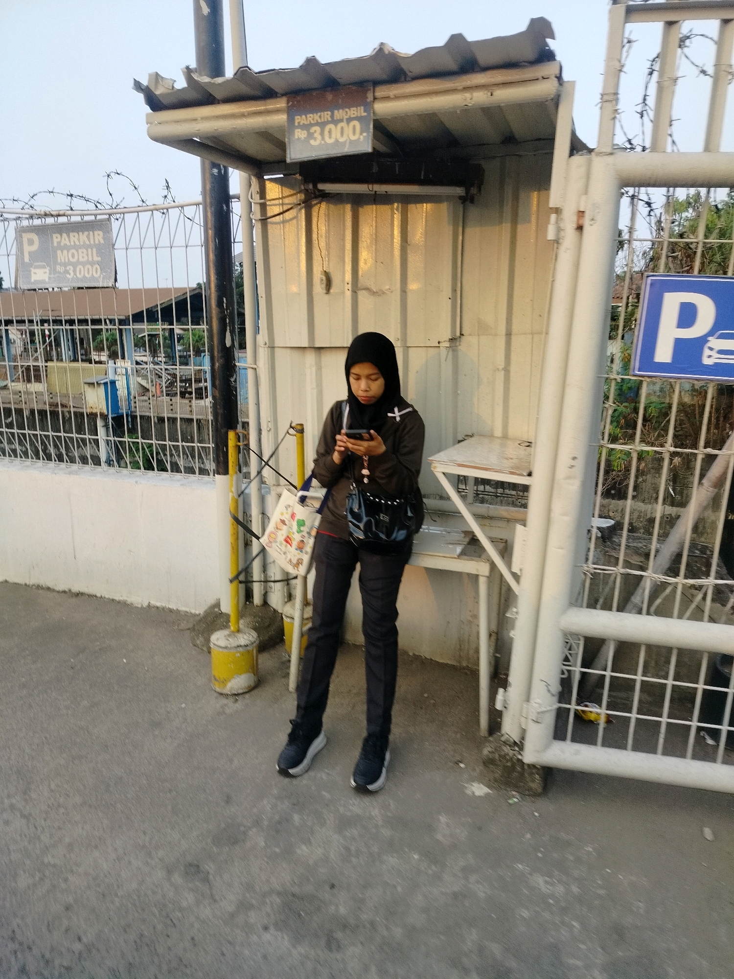 In East Jakarta, Indonesia, a woman wearing a black hijab and casual clothing stands near a parking booth. She is looking down at her smartphone, holding it in both hands. A patterned tote bag hangs from a yellow pole beside her, and a black purse is slung over her shoulder. The parking booth has a corrugated metal roof and a white metal siding. A sign on the booth reads "PARKIR MOBIL Rp 3.000," indicating the car parking fee. Adjacent to the booth is a white metal gate and fence with barbed wire at the top. In the background, a street with a concrete sidewalk and a gray asphalt road is visible, along with some buildings and trees. The overall scene depicts a typical street view in the city, with the woman engaged with her device while waiting.
