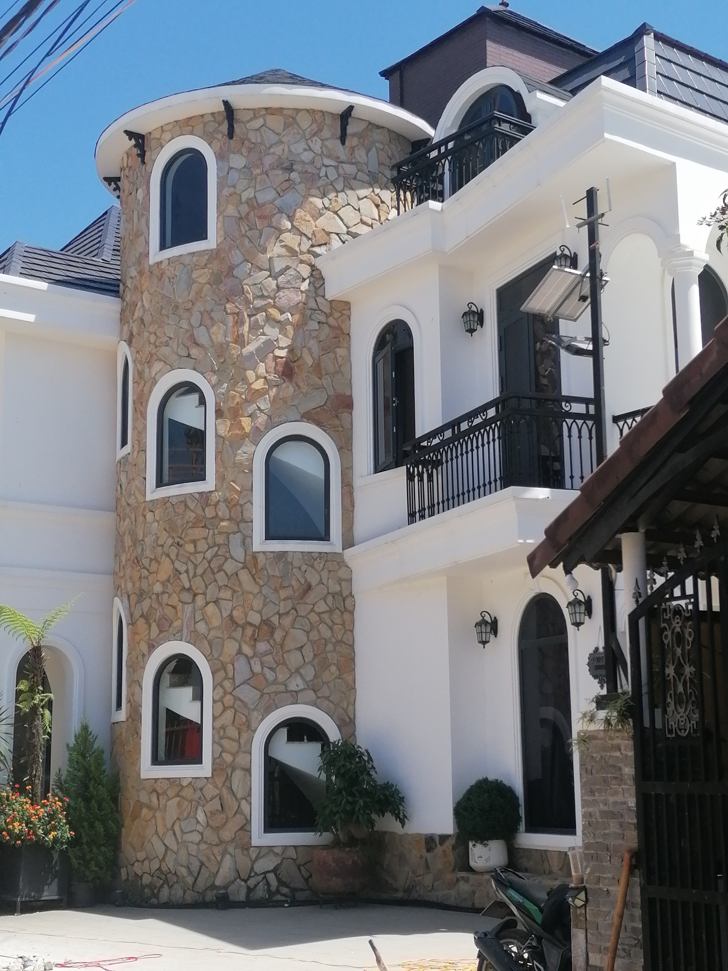 The image depicts a modern building with a distinctive rounded tower constructed of natural stone, integrated with a white stucco facade and dark-tiled roofs. The tower features multiple arched windows with dark frames. The main building showcases a white exterior with arched windows and doors, adorned with black metal railings on balconies and decorative sconce lighting. A solar-powered lighting system is visible on the facade. In the foreground, a motorcycle is parked near a black metal gate. Lush green plants and potted flowers add to the landscaping. The scene is set against a clear blue sky. This building is located in Da Lat, Vietnam, a city known for its French colonial architecture and mountainous terrain.