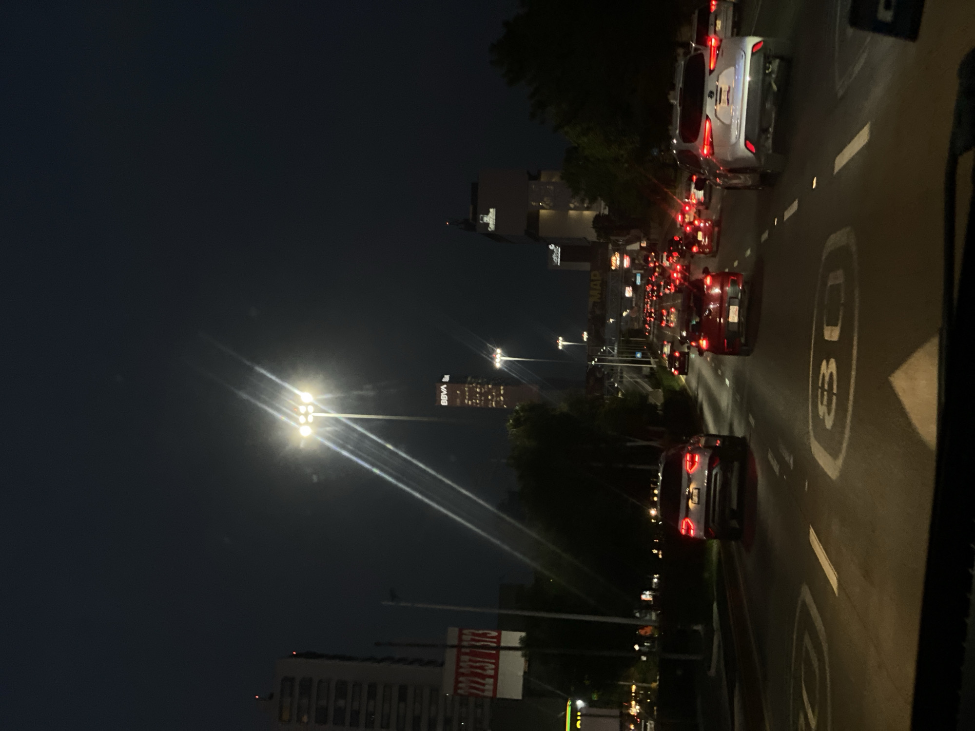 The image depicts a nighttime urban scene with heavy traffic. Cars with lit tail lights are visible on the right side of the image, and streetlights illuminate the road. In the distance, buildings with lit windows rise above the horizon. One building has the letters "BBVA" illuminated on its side. There is a billboard visible on the left side of the road with numbers "777 237 1173" printed on it. A speed limit sign painted on the road shows the number 80. The location is the city of Unknown, Unknown.