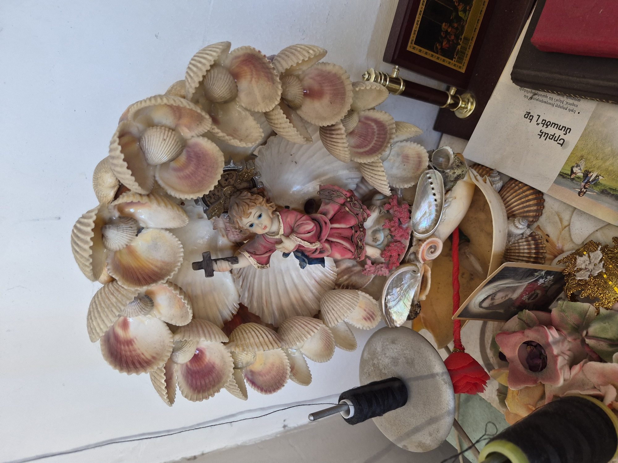 The image captures a detailed, top-down view of a decorative arrangement, likely situated on a flat surface in an indoor setting in Yerevan, Armenia. The central focus is a circular display predominantly made of various seashells, many exhibiting natural pink and purple shades. Nestled within this shell arrangement is a figurine of a child, possibly an angel or baby Jesus, dressed in a pinkish-red garment and holding a small dark cross. The child figurine has light blue eyes.
Around the main shell display, a variety of other items are visible. These include iridescent abalone shell fragments, pieces of red coral, small pink faux flowers, and golden metallic embellishments. A small, vintage-style framed picture, possibly a religious icon or portrait of a woman with a head covering, lies among the objects. Crafting supplies are also present, including spools of black and yellow-green thread, one resting on a flat, light gray stone. In the background, part of a wooden box or frame with a picture and a small wooden and brass telescope-like object can be seen.
A significant detail is a piece of paper with visible text in Armenian. The OCR of this text translates to: "Have you ever wondered? • If everyone wants peace so much, why are there wars?". The overall scene suggests a personal collection of decorative items, possibly with sentimental or religious significance, or a creative crafting space. No direct activity is taking place; the image is a static snapshot of the arrangement.