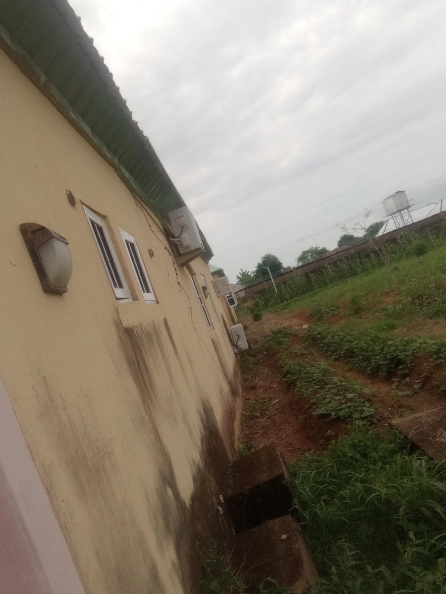 The image depicts an exterior view of a light yellow building with a green corrugated metal roof. The building has several windows with white frames and what appears to be air conditioning units mounted on the exterior wall. There is also a wall-mounted light fixture. The wall shows signs of staining and cracking.
The setting is outdoors, with a grassy area and some cultivated plants visible to the right of the building. In the background, a water tank is mounted on a structure. A fence runs along the property line, separating it from the vegetation and trees beyond. The overall scene suggests a residential or institutional property in a somewhat overgrown or natural environment.
The sky is overcast with clouds, indicating possibly a cloudy day or early morning/late afternoon. There is no clear indication of any specific activity or event taking place. The image is likely a photograph taken during daylight hours. The location context is Jalingo, Nigeria, which aligns with the appearance of the vegetation and building style. No visible text is present in the image.