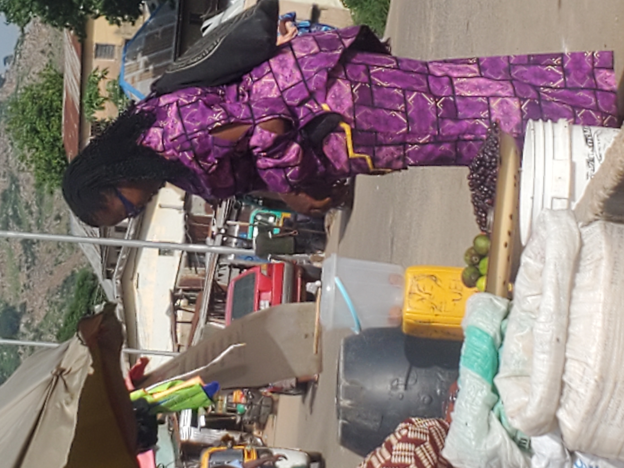 A person, wearing a purple and gold patterned dress and a black headwrap, stands in a busy outdoor market. The person appears to be a vendor, with various goods arranged around them. In the foreground, there are bunches of dark berries and what appear to be kiwis. Behind the person, there are plastic containers, bags of what could be produce or other market goods, and what looks like a red food cart. In the background, buildings and a hillside are visible. The lighting suggests it is daytime. The scene is likely in Gwong, Nigeria, given the context.