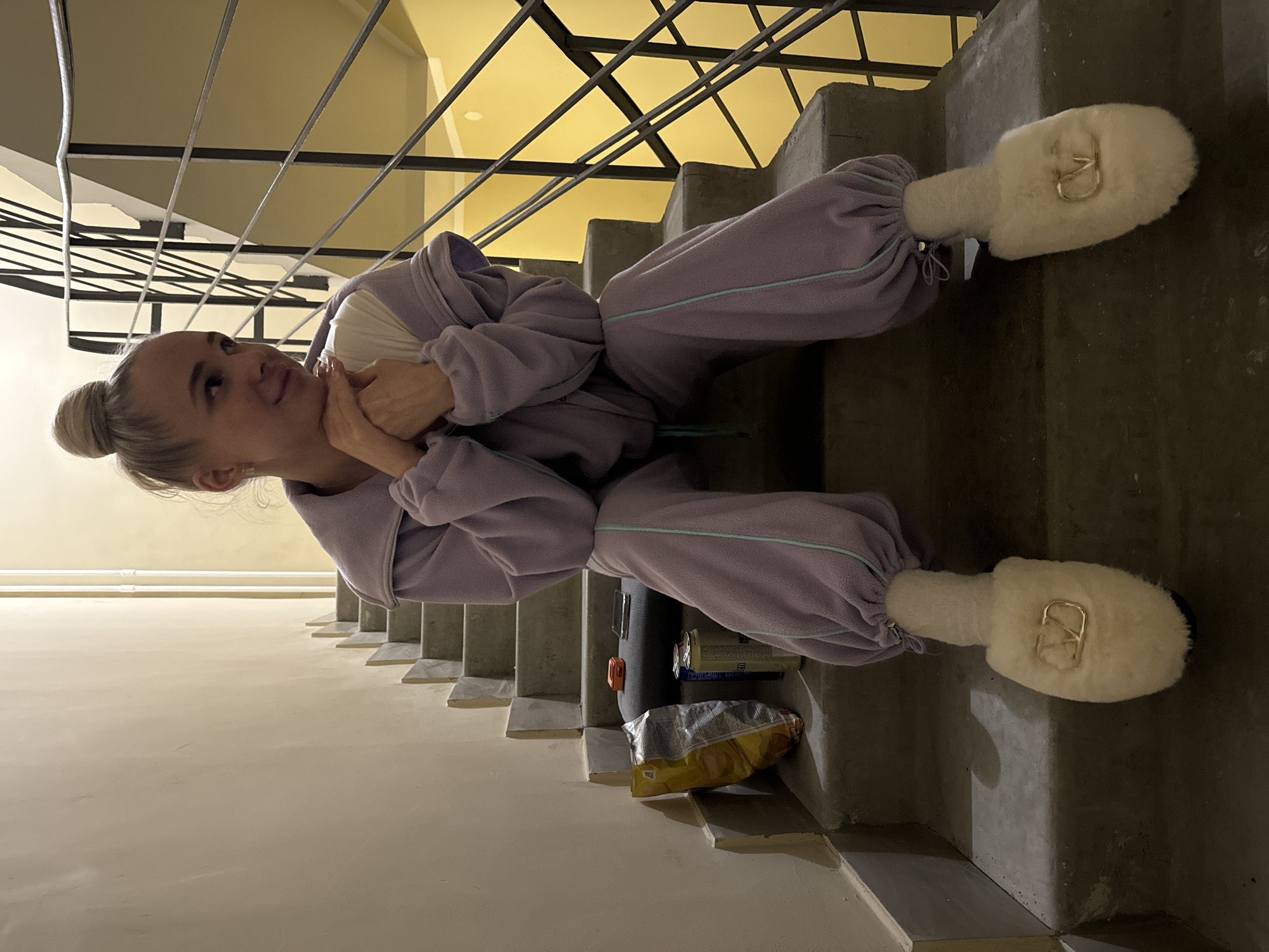 A Caucasian woman with fair skin and blonde hair pulled back into a bun sits sideways on a concrete staircase, her head turned to the left. She wears a light purple tracksuit with turquoise trim and large, furry white slippers with gold embellishments. Her hands are clasped together under her chin.
To the left of the woman are beige walls and black metal stair railings. A bag of chips, a can of something, and a black object rest on the steps near her.
The photo was taken in what looks like an apartment building. The stairs are in Akulovo (Gp Kubinka), Russia.