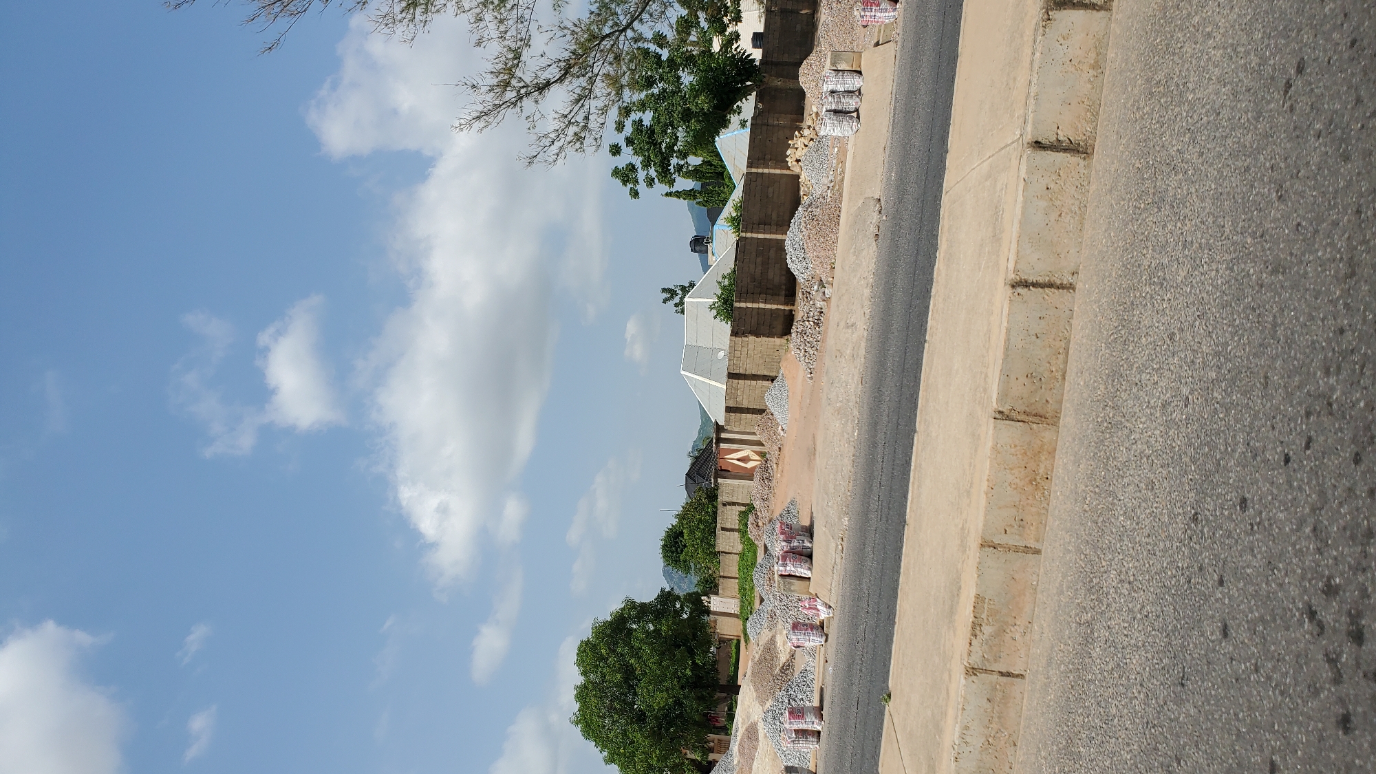 The image depicts an outdoor scene on a bright, cloudy day, likely in Jalingo, Nigeria. The perspective is from a street level, looking upwards towards a mix of residential structures and construction materials.
The foreground is dominated by an asphalt road and a concrete curb, suggesting an urban or semi-urban environment. Along the side of the road, there are stacks of construction materials, primarily bags of cement and piles of gravel. These are positioned near a brick wall that delineates a property.
Beyond the wall, several houses are visible. Some have pitched roofs covered with what appears to be corrugated metal, while others have flatter designs. The architecture suggests a developing area, with visible construction and building activity. Green trees are scattered throughout the scene, indicating a potentially warm climate. In the background, hills or mountains can be seen under the hazy sky.
The sky is a prominent feature, filled with large, fluffy white clouds against a bright blue backdrop. The lighting suggests it is daytime, possibly late morning or early afternoon, given the intensity of the light. There is no visible text in the image. The scene conveys a sense of ongoing development and daily life in a town or city.