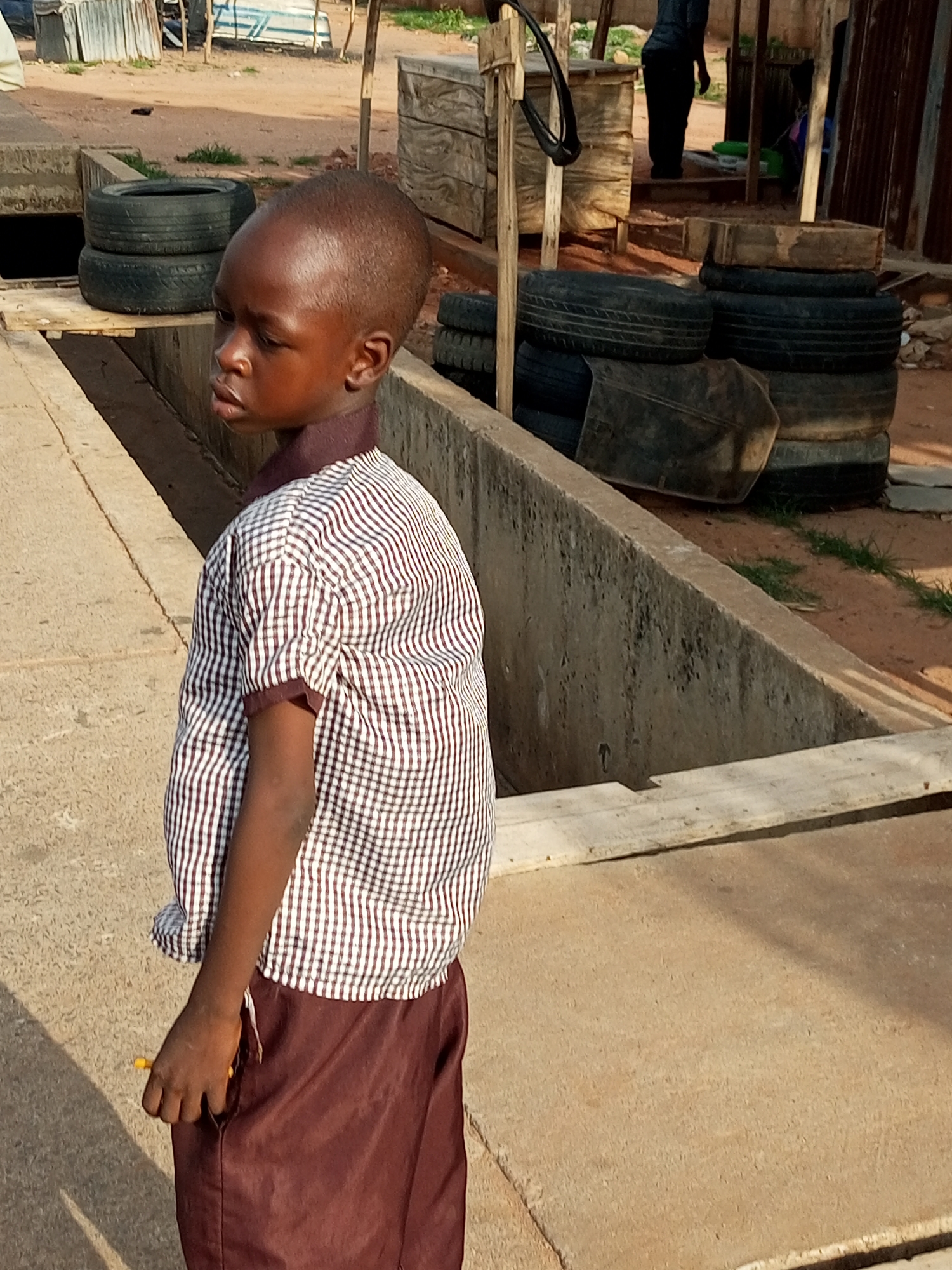 A young boy stands in a side profile to the right, facing left, with his head turned further left, appearing to look downwards. He wears a collared, short-sleeved shirt with a brown and white checkered pattern and maroon bottoms. His right hand is visible at his side, holding a yellow object. The boy is positioned outdoors on a paved surface, possibly a walkway or road, with a concrete structure extending into the foreground. Behind him, to his left, are several stacked tires. In the background, various structures and objects are visible, including what appear to be wooden crates, corrugated metal sheeting, and additional tires. The ground is dusty and sandy. The scene suggests an outdoor setting in a possibly rural or semi-industrial area. The location is identified as Wuro Dole, Nigeria.