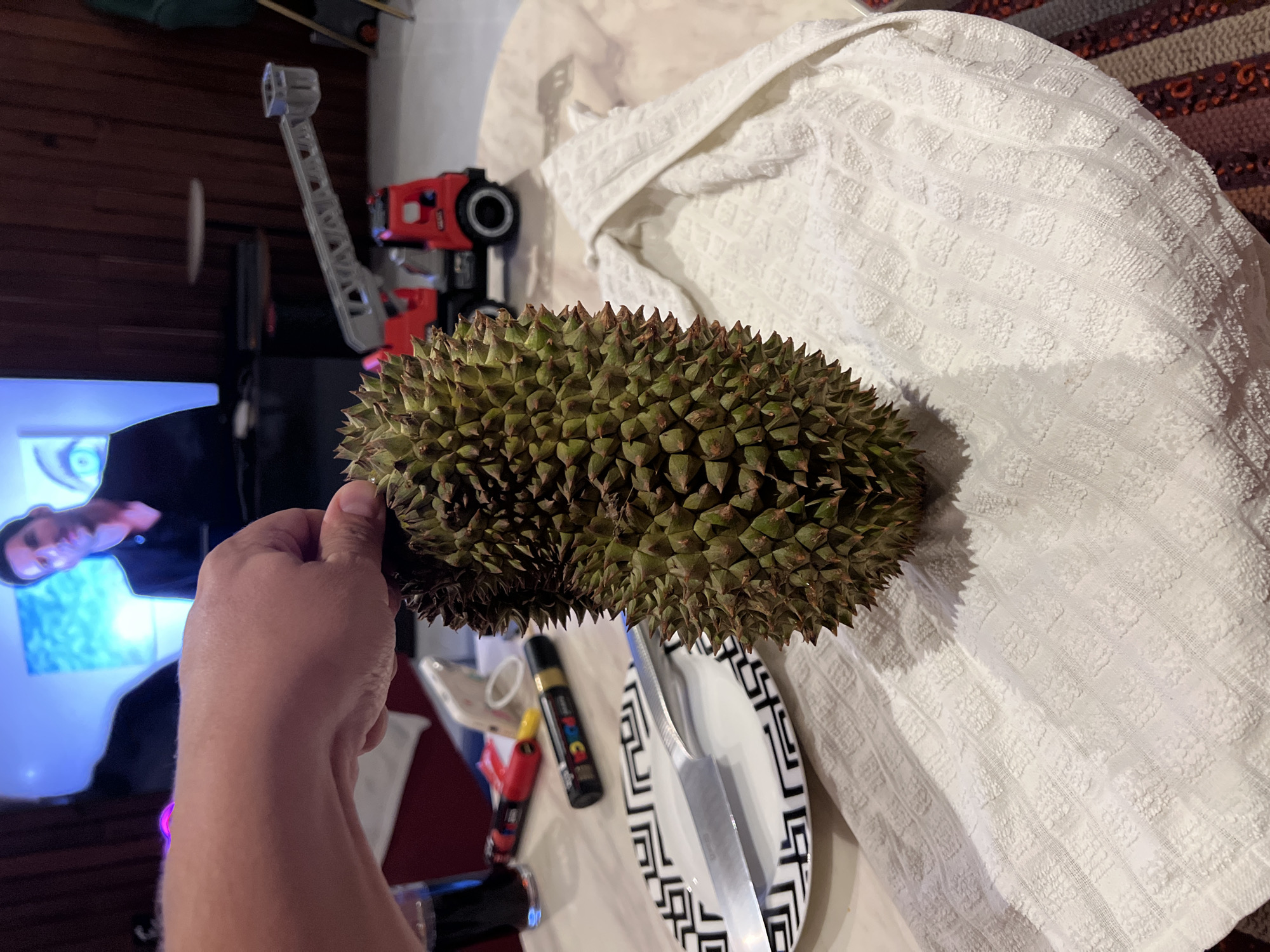 The media file shows a close-up view of a durian fruit being held in a person's hand. The durian is large and spiky, with a green rind. The hand holding the fruit is light-skinned. In the background, there is a television screen displaying a person's face, a red and black toy vehicle with a crane, a white textured cloth, and a plate with a knife and some markers. The setting appears to be indoors, possibly a dining area or living room, with a striped rug visible in the upper right corner. The lighting suggests it could be evening or nighttime, given the illuminated television screen and the indoor environment. The overall mood is neutral, with no strong emotions conveyed. There is some text visible on the markers: "POSCA" and "B".