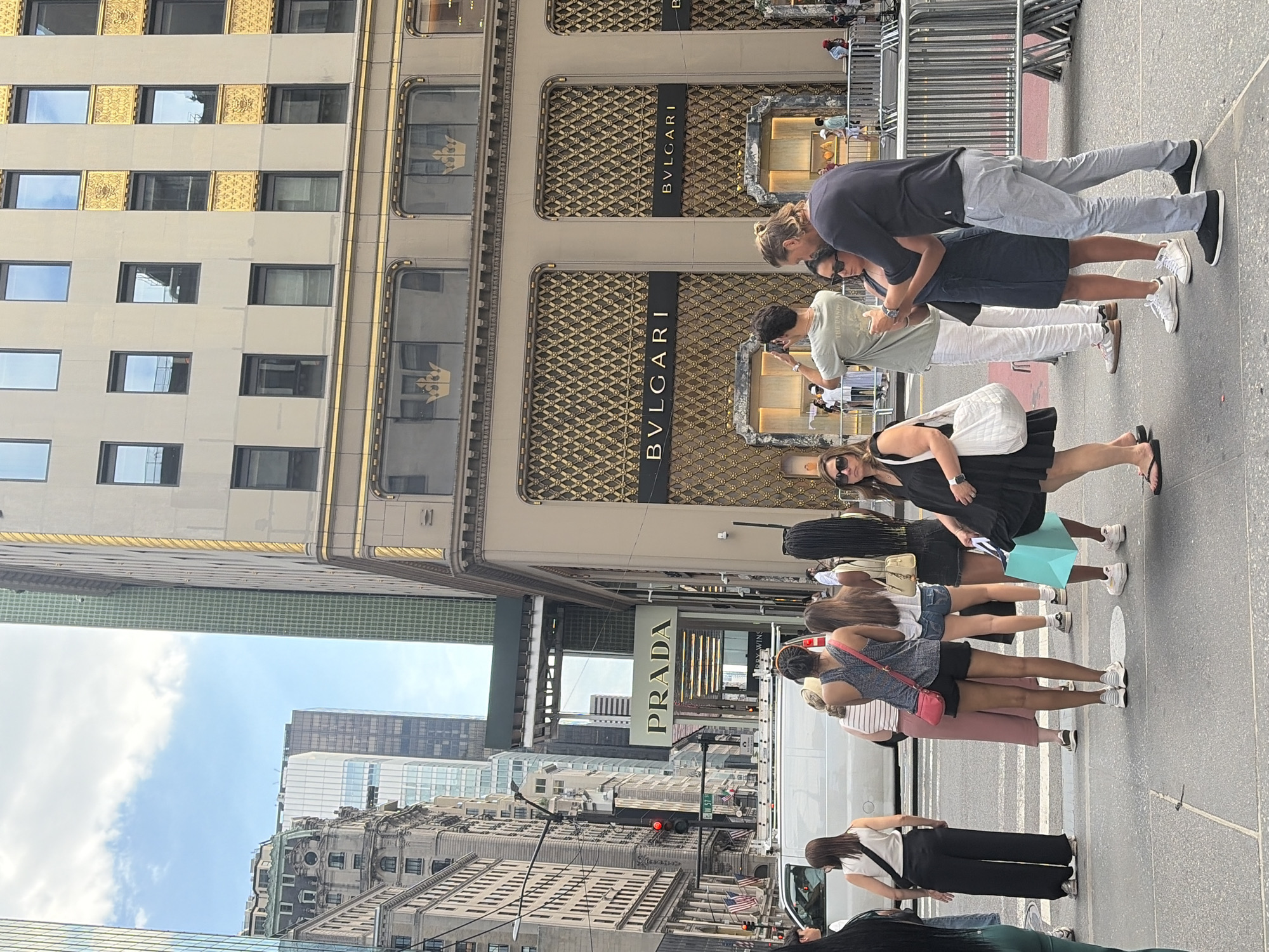 This media file captures a bustling street scene in New York City. The foreground is dominated by a group of people, appearing to be tourists or shoppers, standing on a sidewalk. They are casually dressed, suggesting a daytime activity under fair weather conditions, with a bright sky visible in the background.
The backdrop features impressive architecture, indicative of a commercial district. A prominent building with a gold-patterned facade and the "BVLGARI" logo suggests a luxury retail area. Further down the street, the "PRADA" sign on another building reinforces this impression. The presence of multiple people and the general urban environment point towards a lively, perhaps even crowded, atmosphere.
The people in the foreground are diverse in age and appearance. Some are looking at their phones, while others are engaged in conversation or observing the surroundings. Their stances and expressions are neutral, conveying a sense of everyday activity rather than a specific event. A white van and other vehicles are visible, indicating traffic and the typical flow of a city street. The overall impression is a snapshot of urban life in a well-known shopping and entertainment district.