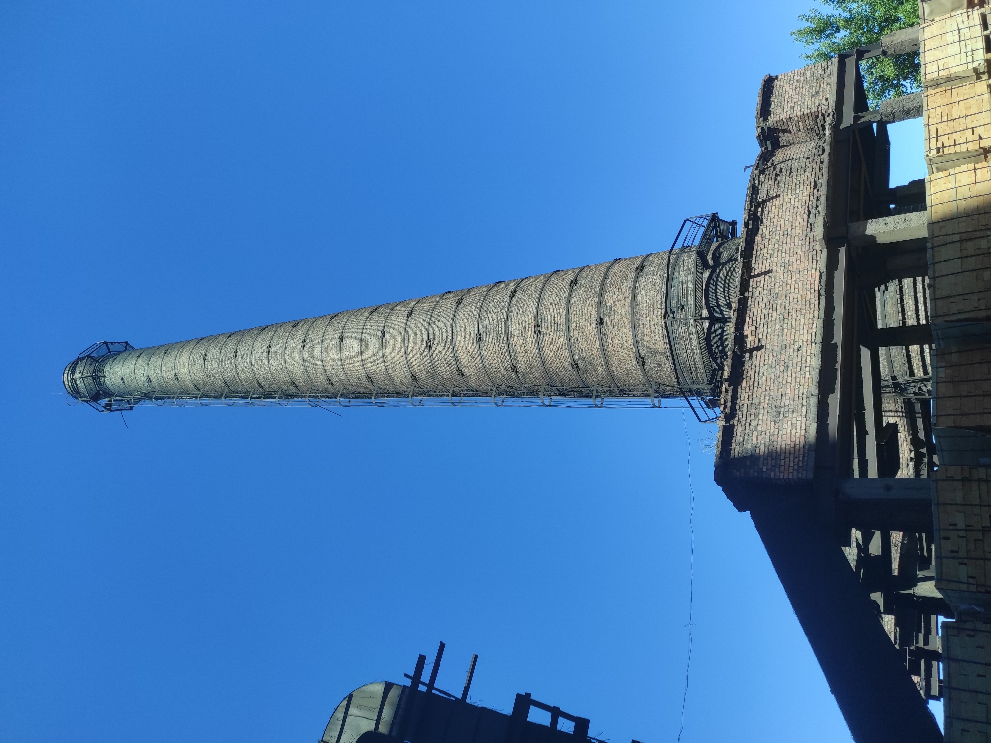 In the city of Kyzyl, Kazakhstan, a tall, weathered brick smokestack rises against a clear, vibrant blue sky. The smokestack, constructed from many courses of reddish-brown bricks, shows signs of age and disuse. A metal ladder, affixed to the exterior, ascends along its entire height, culminating in a caged platform at the very top. The base of the smokestack is anchored to a brick structure with a damaged, crumbling facade, supported by a framework of exposed metal beams and concrete pillars. To the left of the smokestack, a dark, angular structure, possibly part of an industrial building, looms in the foreground, its framework also partially visible. In the lower portion of the image, stacks of rectangular, light-colored objects, possibly bricks or building materials, are neatly arranged. A few green trees are visible in the background on the right, adding a touch of nature to the industrial landscape. The scene appears to be a snapshot of a disused industrial site under a bright, sunny day.