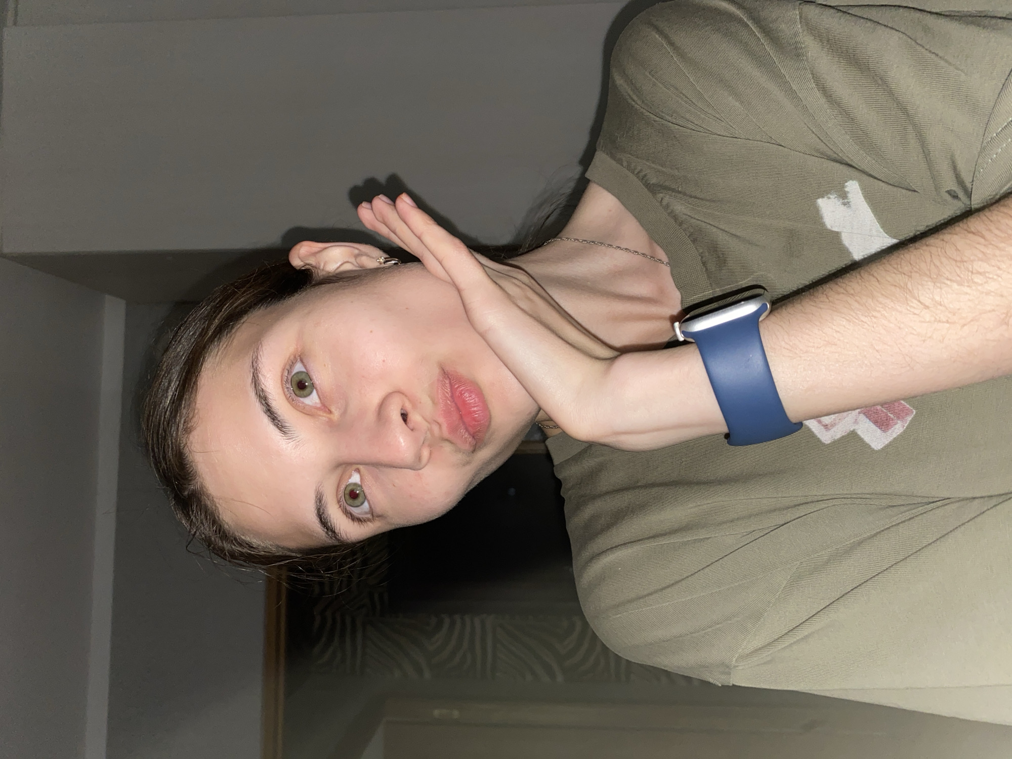 The image shows a young woman with light skin, brown hair, and green eyes, wearing an olive-green t-shirt and a blue smartwatch. She is holding her right hand to her face, with her lips pursed as if blowing a kiss. The background is dimly lit and appears to be an indoor setting, possibly a bedroom or living room, with a wall and some patterned wallpaper visible. The lighting suggests it might be nighttime or a dimly lit interior. There is no discernible indication of the location being Votkinsk, Russia.