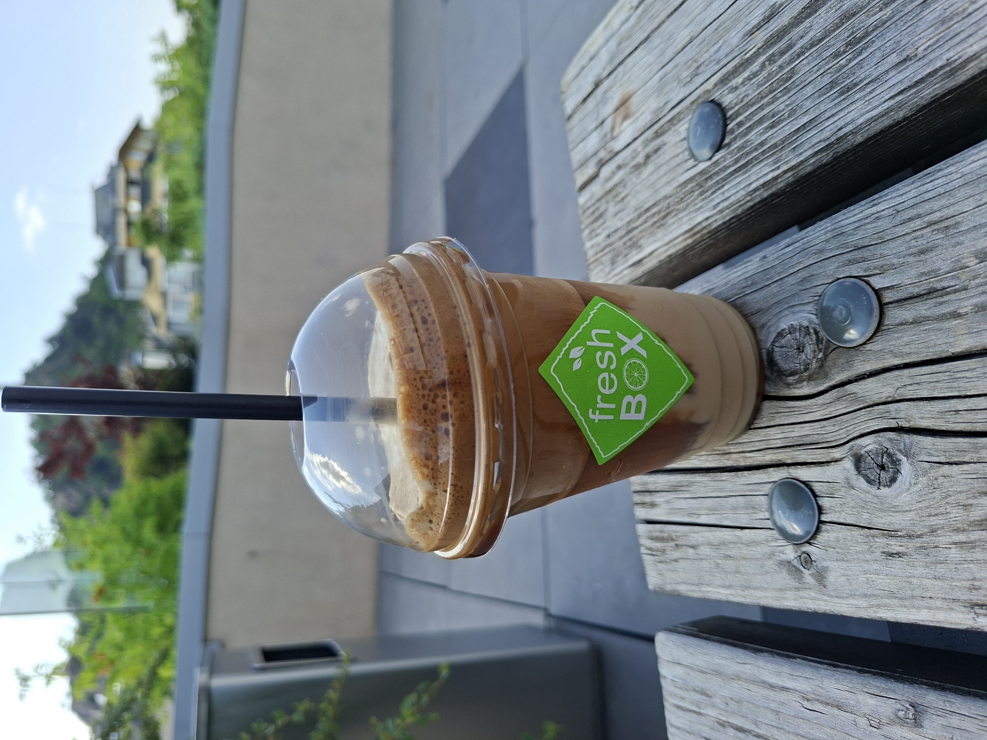 The image displays a clear plastic cup filled with a light brown, foamy beverage, likely an iced coffee or frappe, topped with a domed lid and a black straw inserted into it. A bright green, square-shaped sticker is prominently affixed to the side of the cup, featuring white text that reads "fresh BOX." Above "fresh," a small green leaf icon is visible, and next to "BOX," there's a graphic of a green lime or lemon slice.
The cup rests on a sturdy, weathered wooden bench, characterized by its visible grain, knots, and dark circular metal fasteners (possibly bolts or rivets) embedded in its surface. The texture of the wood suggests age and outdoor exposure.
In the blurred background, a bright daytime scene unfolds under a light blue sky with a hint of a white cloud. Distant buildings, possibly residential or commercial structures, are nestled on a verdant hill covered with lush green trees and foliage. Closer to the foreground, but still out of focus, are elements of modern architecture, possibly a glass barrier or a light-colored building, alongside more greenery. The overall setting appears to be an outdoor urban or park environment, suggesting a pleasant day in Plovdiv, Bulgaria. The image is oriented vertically (rotated 90 degrees clockwise), making the horizon appear on the left side of the frame.