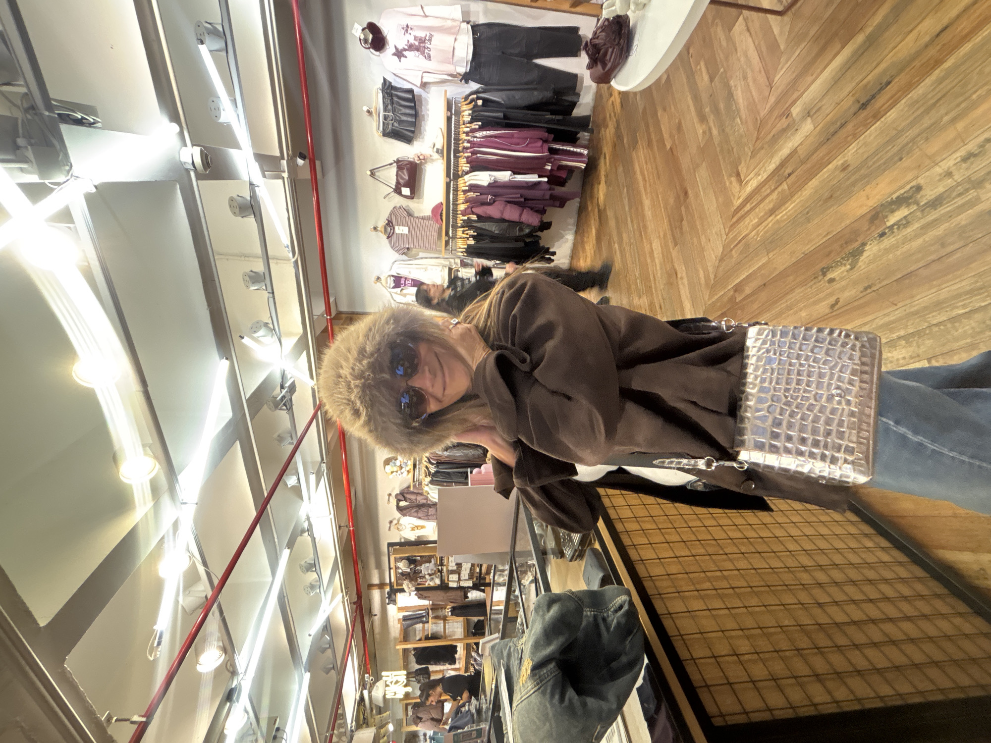 A person wearing a fur hat and sunglasses is posing for a photo inside a clothing store. They are wearing a brown jacket and carrying a silver purse. The store is filled with racks of clothing and shelves of merchandise. The location appears to be a clothing store based on the presence of racks of clothing, and shelves of merchandise in the background. It is difficult to verify the exact location in Buenos Aires, Argentina without more context or visual cues from the environment.